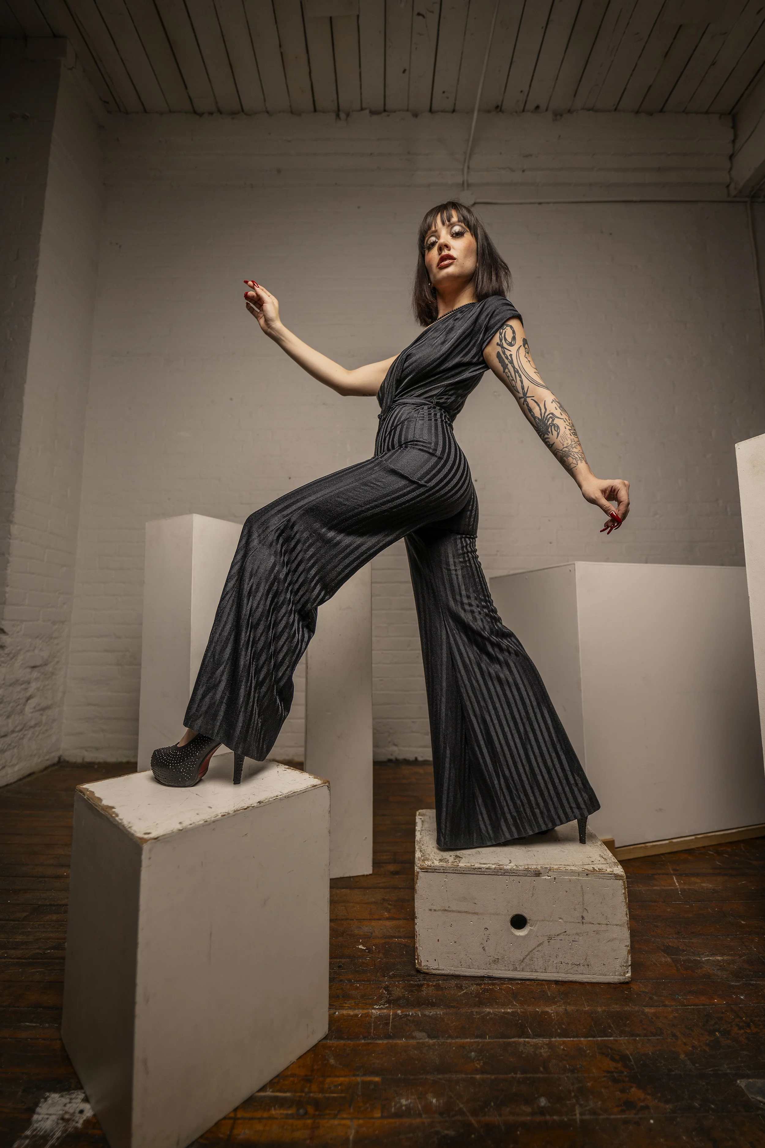 A woman with short dark hair, wearing a black striped jumpsuit and high heels, posing on white boxes in a minimalist room with a white brick wall and wooden floor.