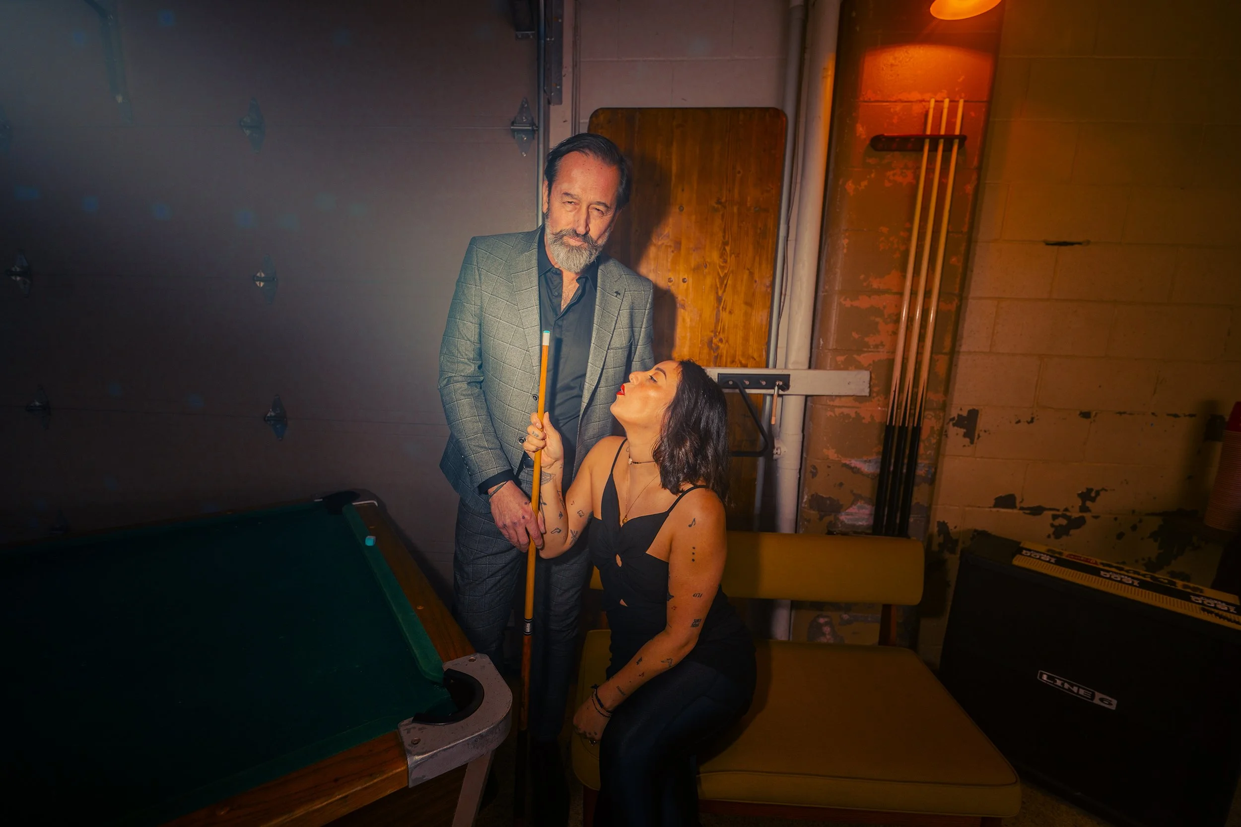 A man and a woman are playing billiards in a dimly lit room. The man is holding a cue stick and looking down at the woman, who is seated and holding her own cue stick. The room has a pool table, a brick wall, and some pool cues in the background.