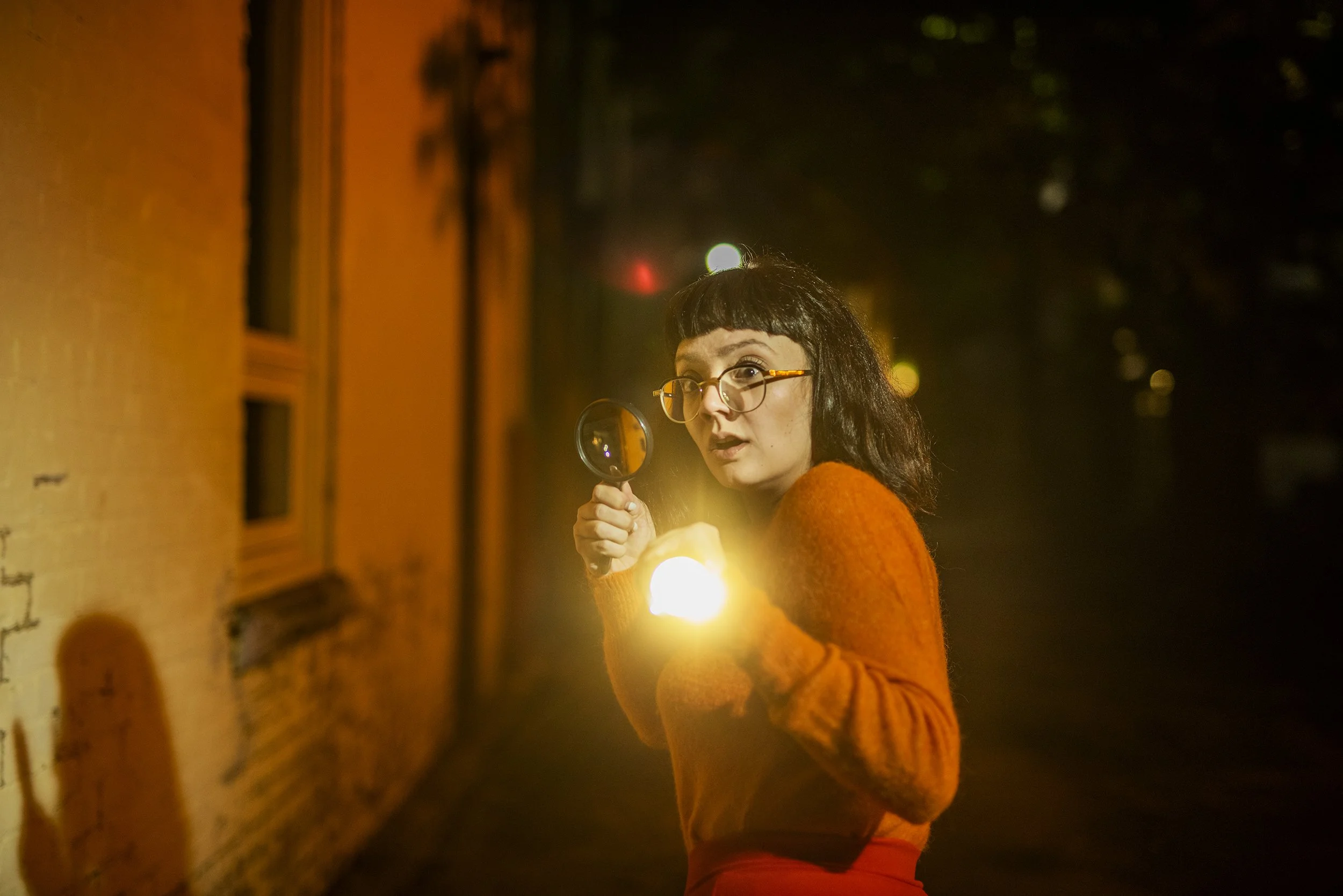 A woman with dark hair, glasses, and bangs, dressed in an orange sweater, holding a magnifying glass and looking curious at night in an urban setting with yellow-orange lighting and blurred background.