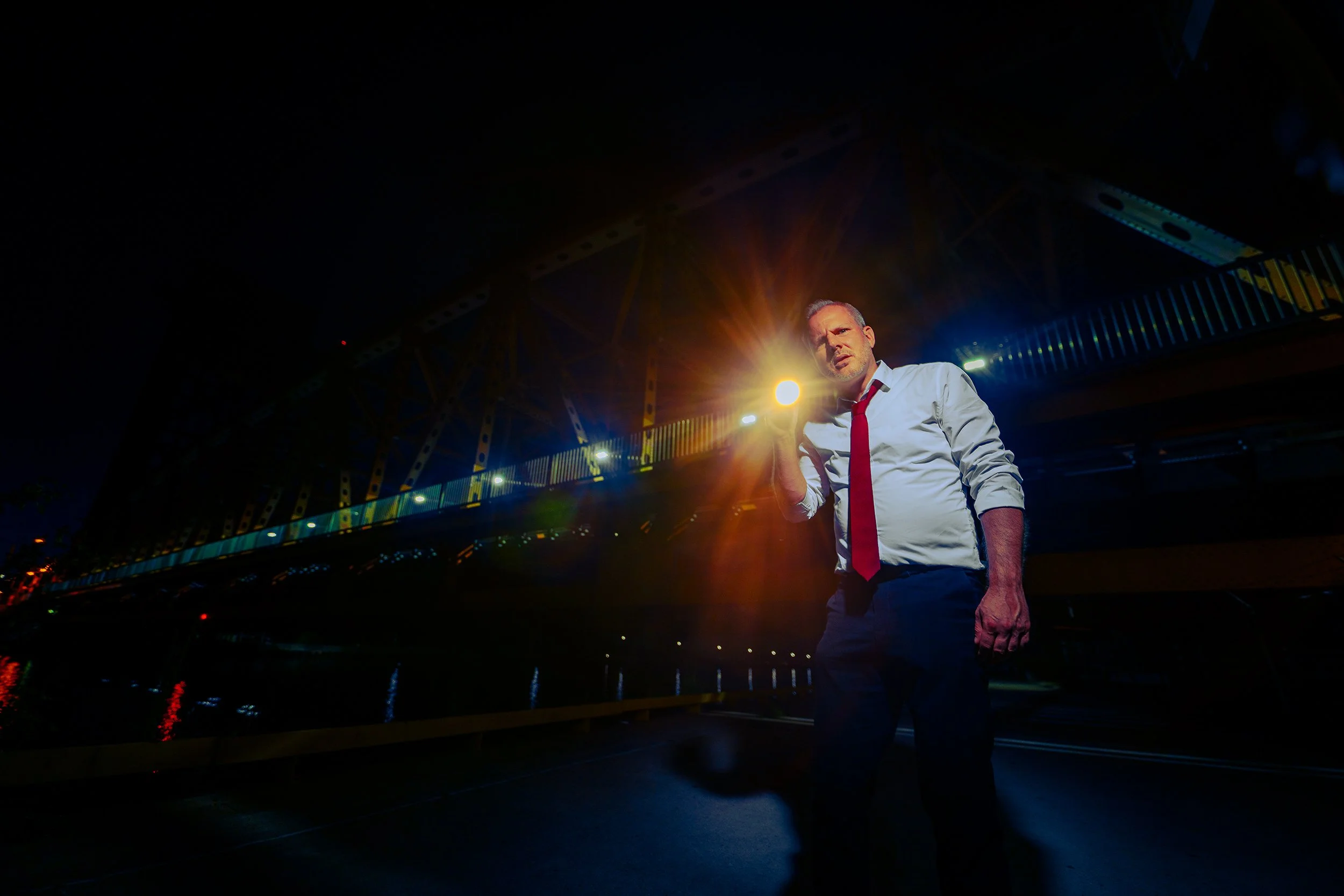 A man in a white shirt and red tie stands under a bridge at night with bright lights and reflections on the water behind him.