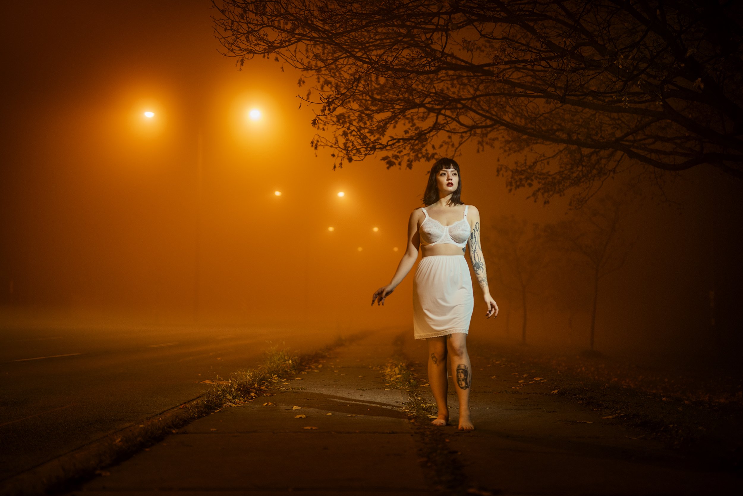 A woman with tattoos on her arms and legs, wearing a white bra and skirt, walking barefoot at night on a foggy street with orange streetlights and leafless trees overhead.