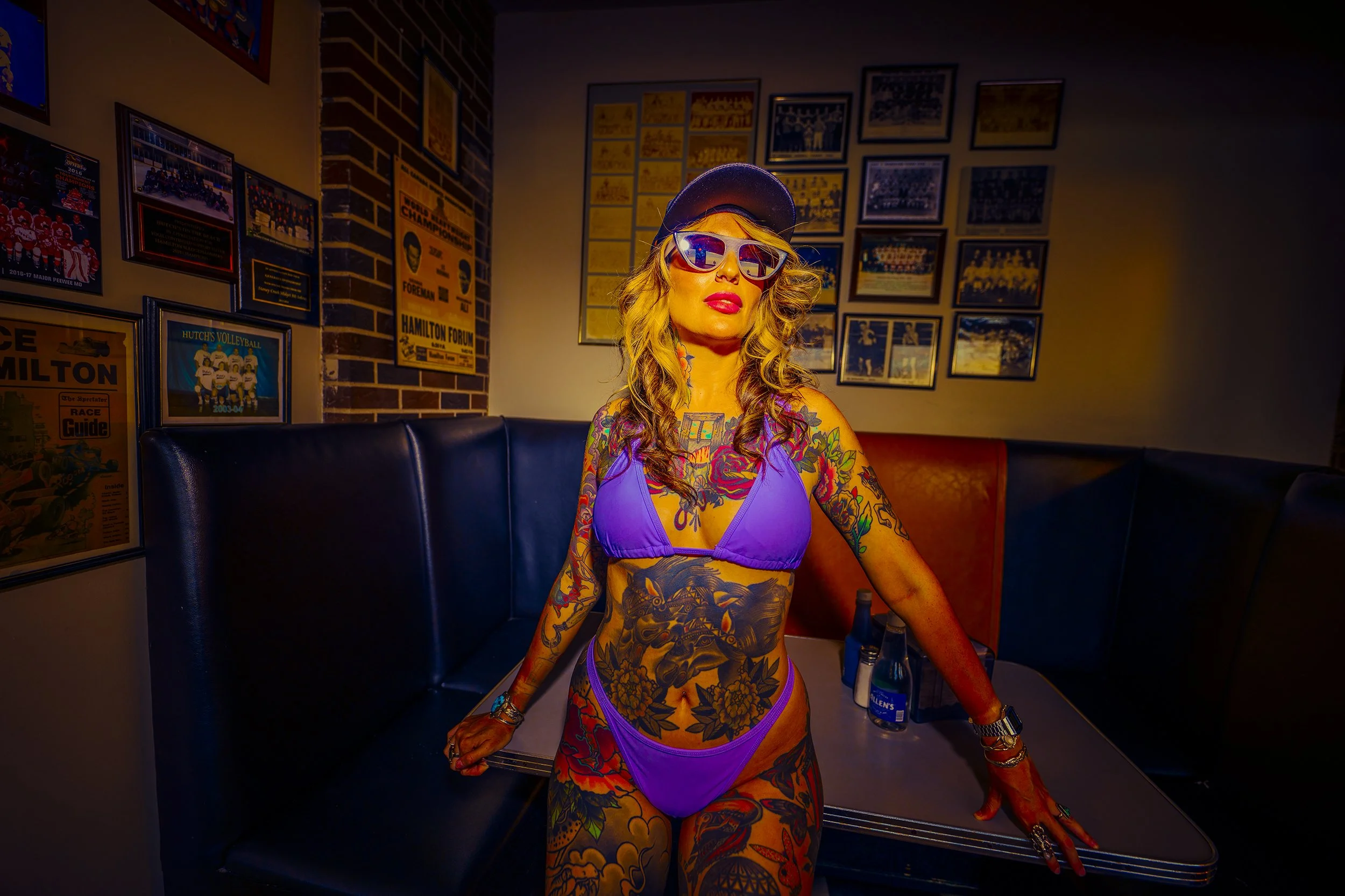 A tattooed woman wearing purple bikini, sunglasses, and a cap, standing in a vintage-style diner with framed sports memorabilia on the walls.