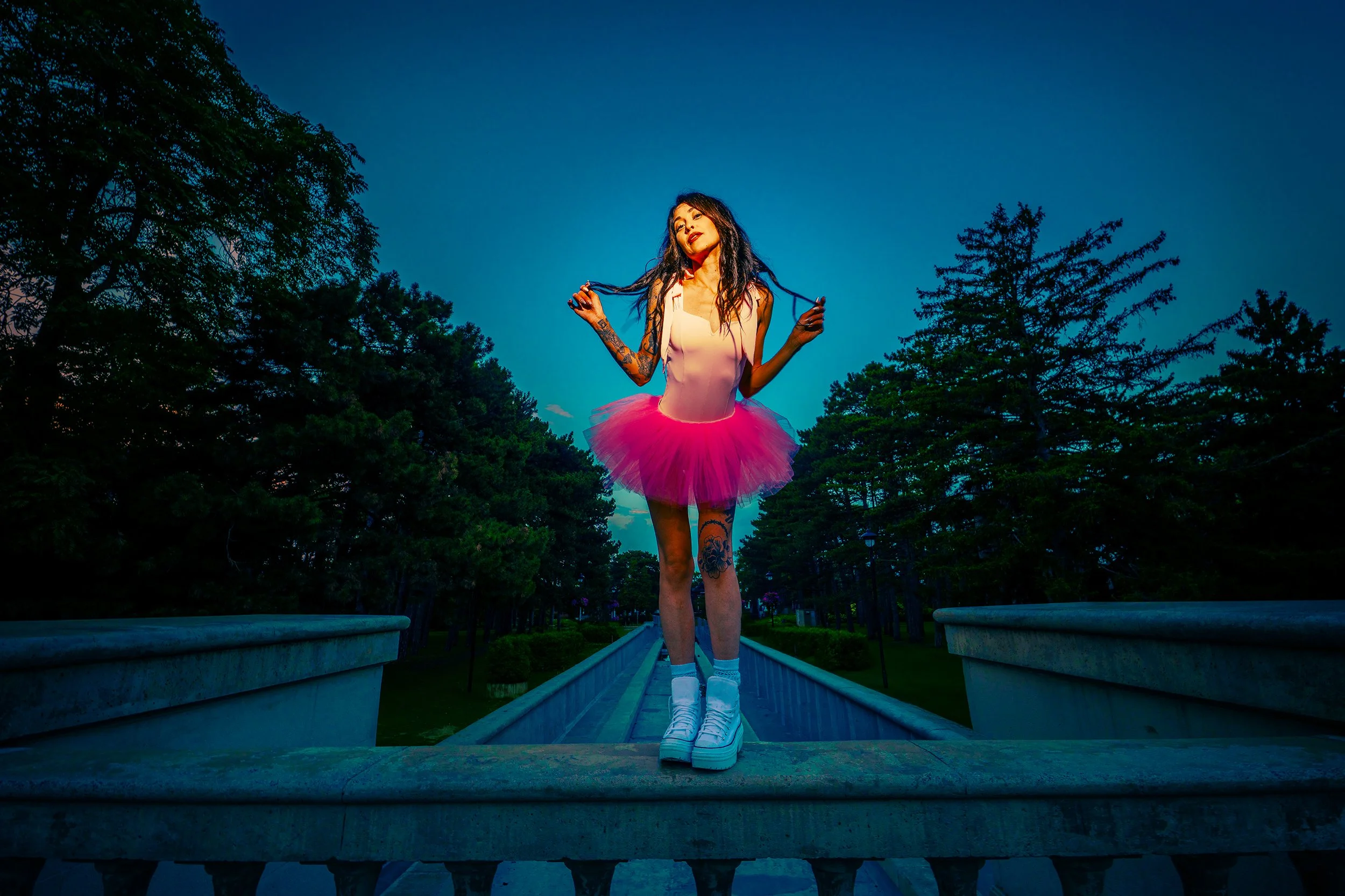 A woman in a pink tutu skirt, white crop top, and platform sneakers standing on a bridge in a park at dusk, holding her long dark hair.