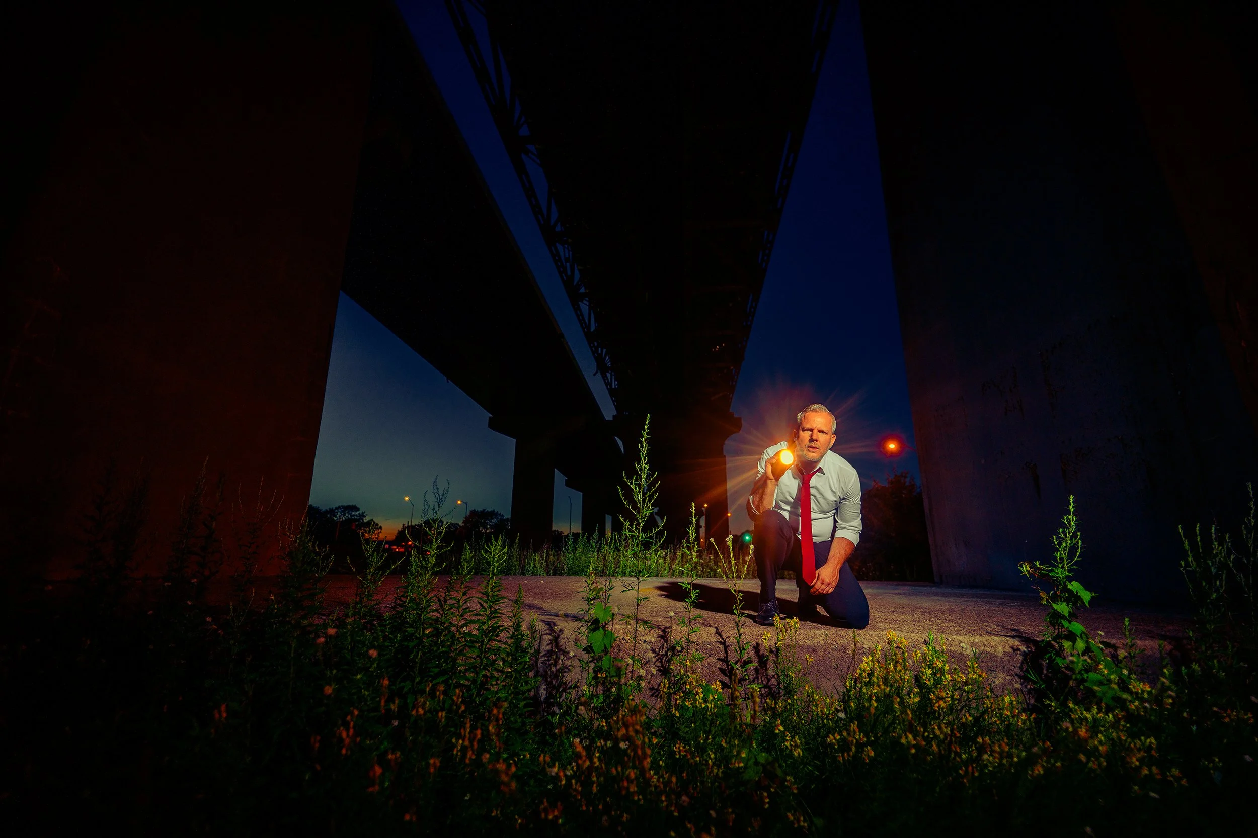 A man in business attire kneels under a bridge at dusk, holding a flashlight, with the bridge's structure silhouetted against the evening sky.