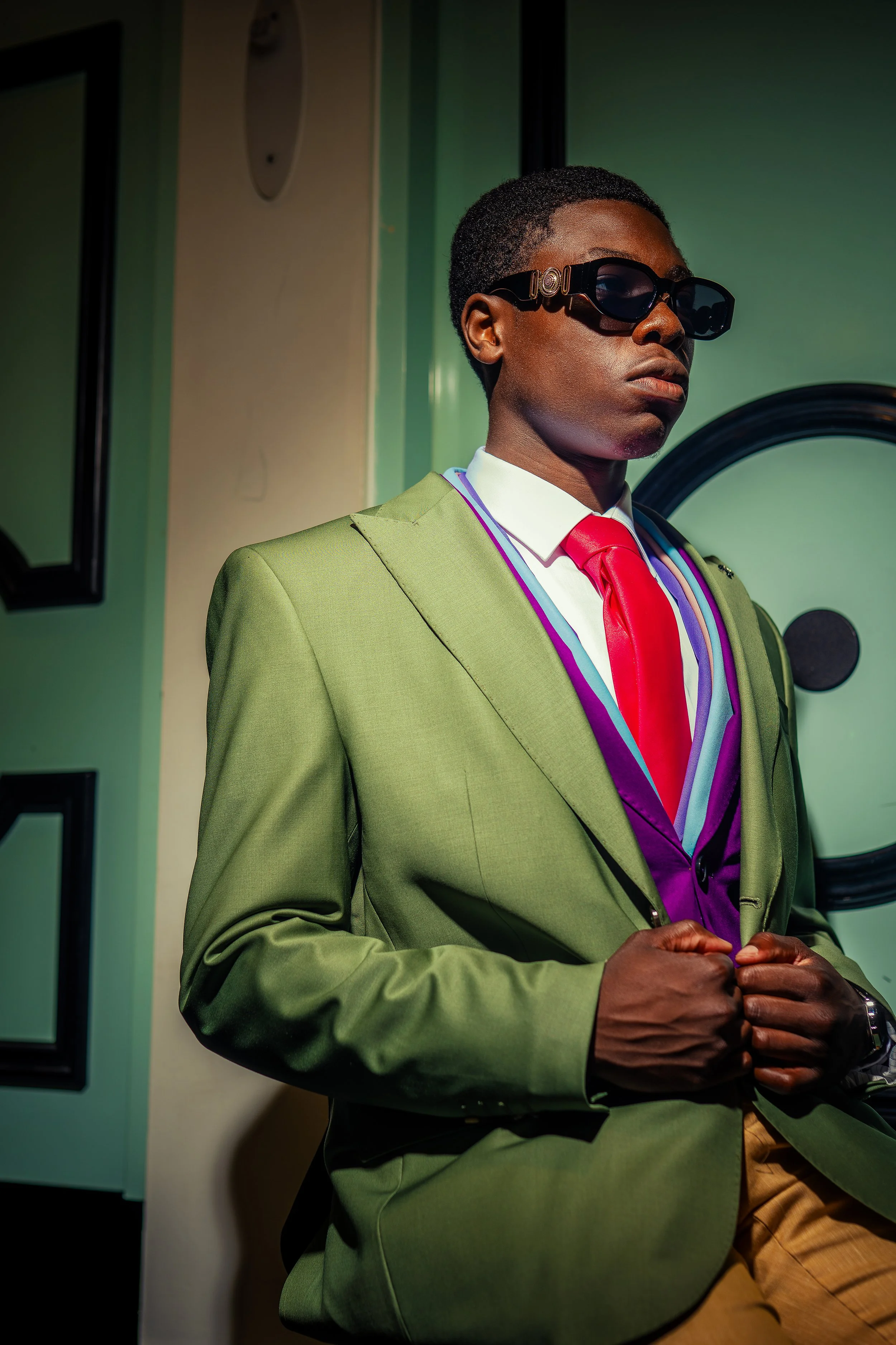 A man wearing a lime green suit, red tie, white shirt, and tan pants, with dark sunglasses, standing indoors against a yellow wall and a green wall with black picture frames.