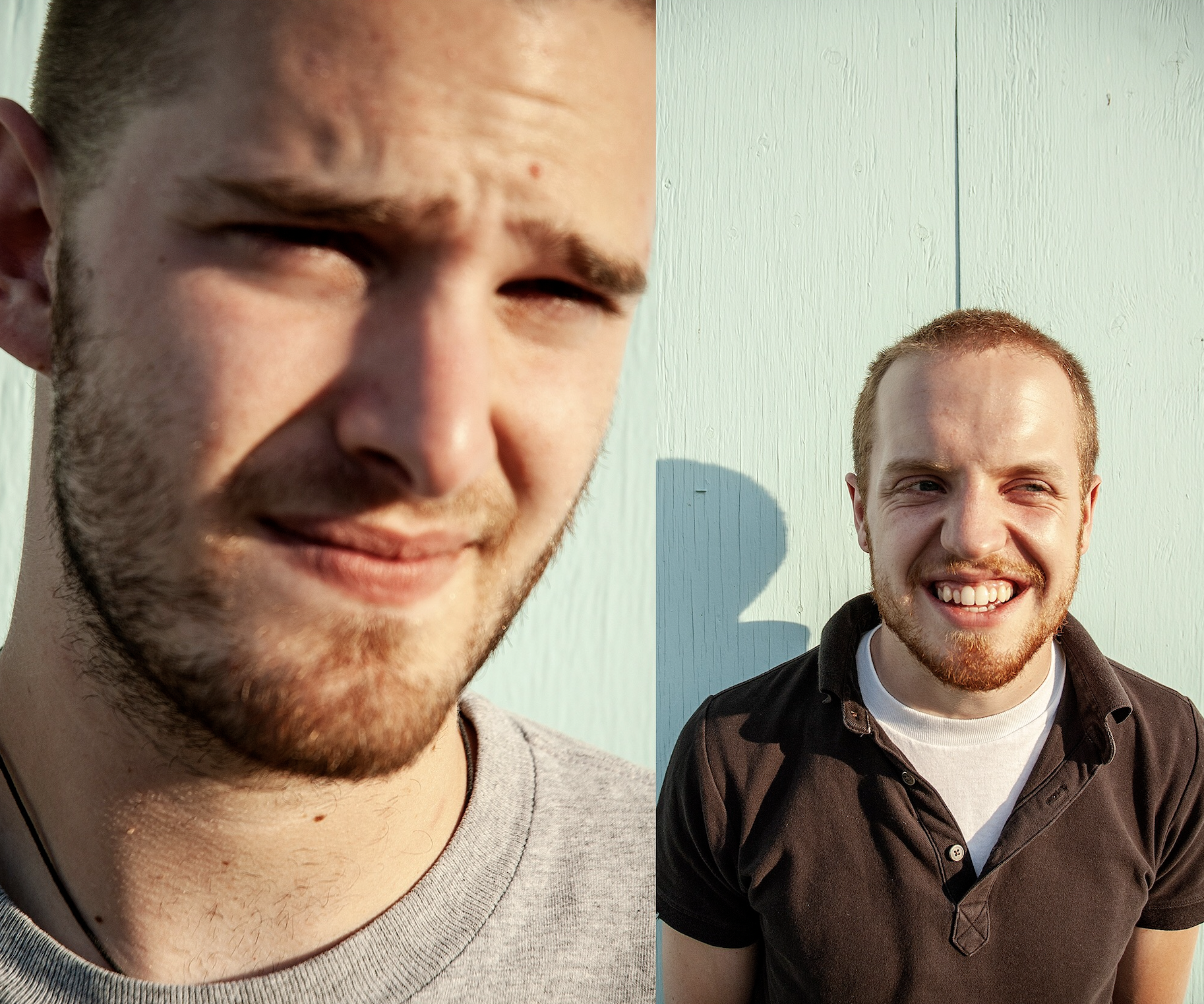 Two young men smiling outdoors against a pale green wall, sunlight casting shadows. The man on the left has a beard and is wearing a light gray shirt, and the man on the right has short red hair, a beard, and is dressed in a black shirt over a white 
