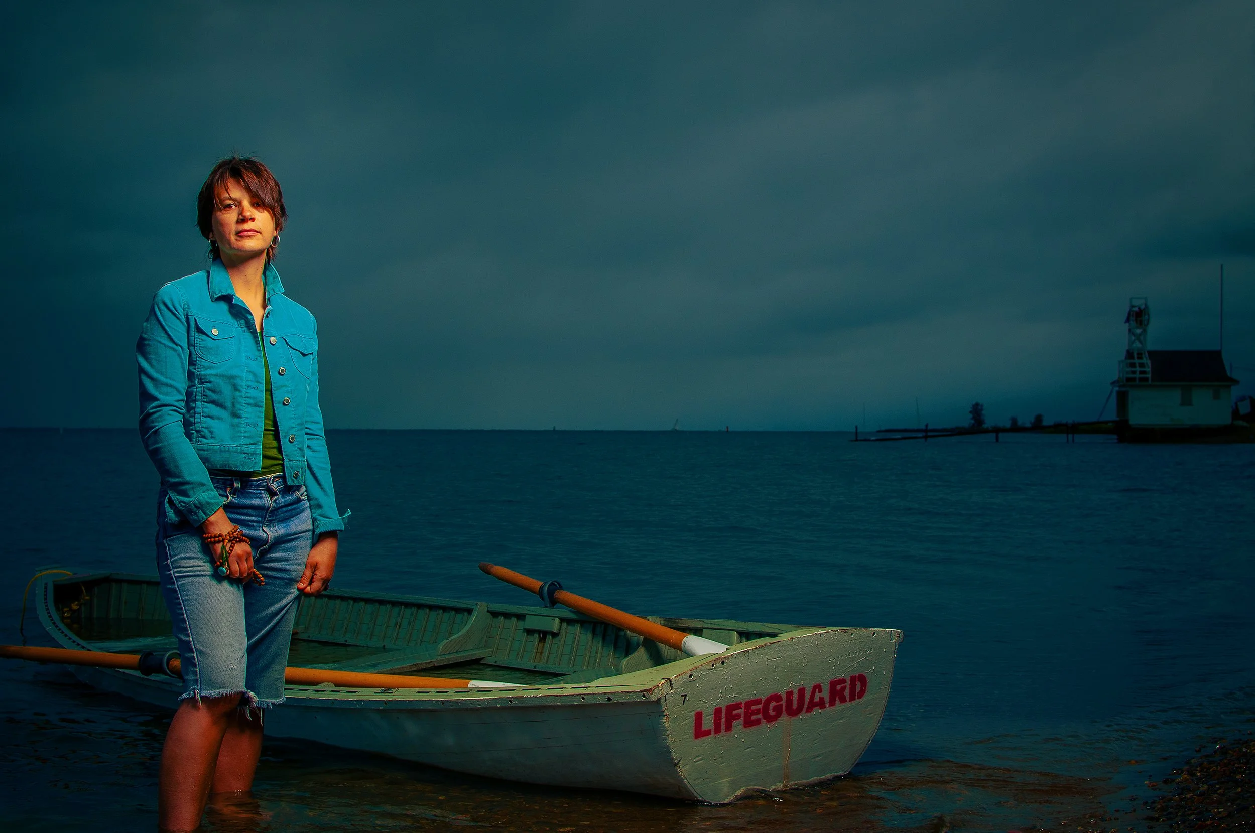 A woman with short dark hair and wearing a turquoise jacket and denim shorts standing knee-deep in water next to a boat labeled 'LIFEGUARD' near a shoreline with a dark cloudy sky and a lighthouse in the distance.