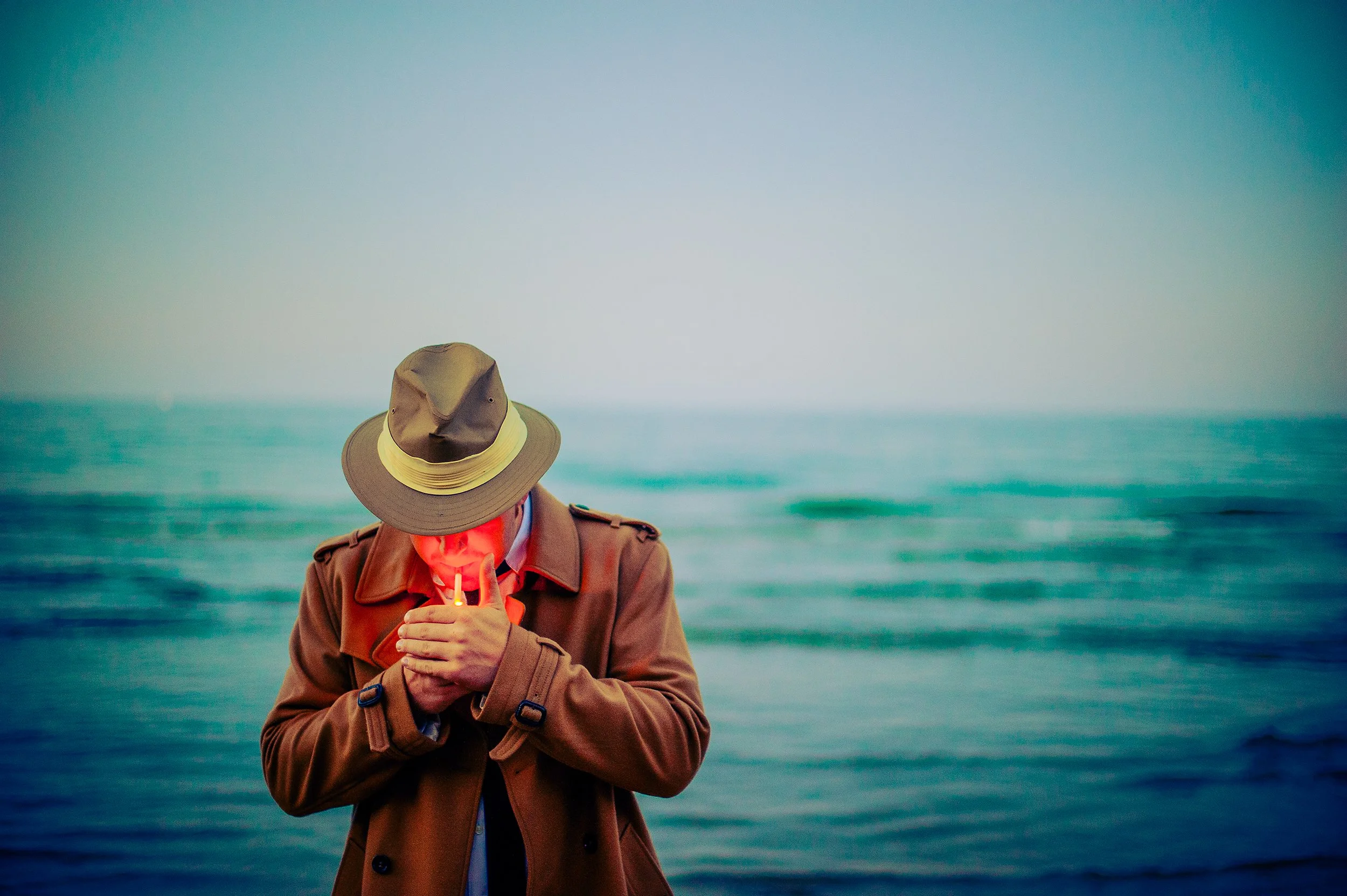 Person in a brown coat and wide-brim hat holding a lit candle at the beach.