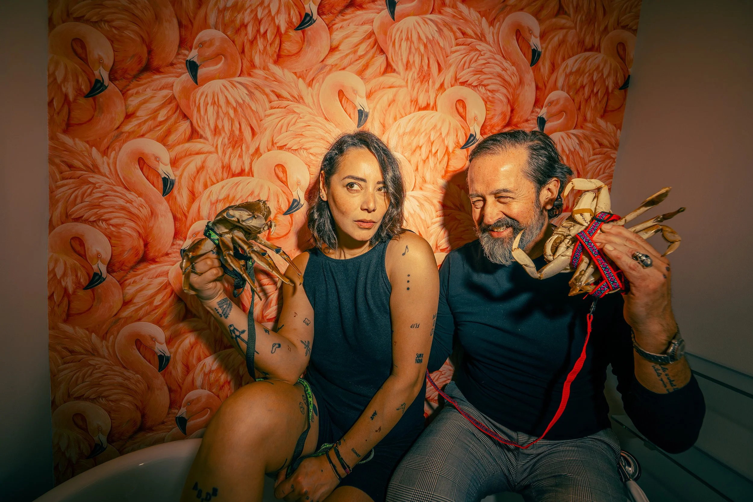 A woman with tattoos and shoulder-length dark hair, and a man with tattoos and a beard, sitting in front of a background with illustrated pink flamingos. They are holding large, realistic crab claw props.