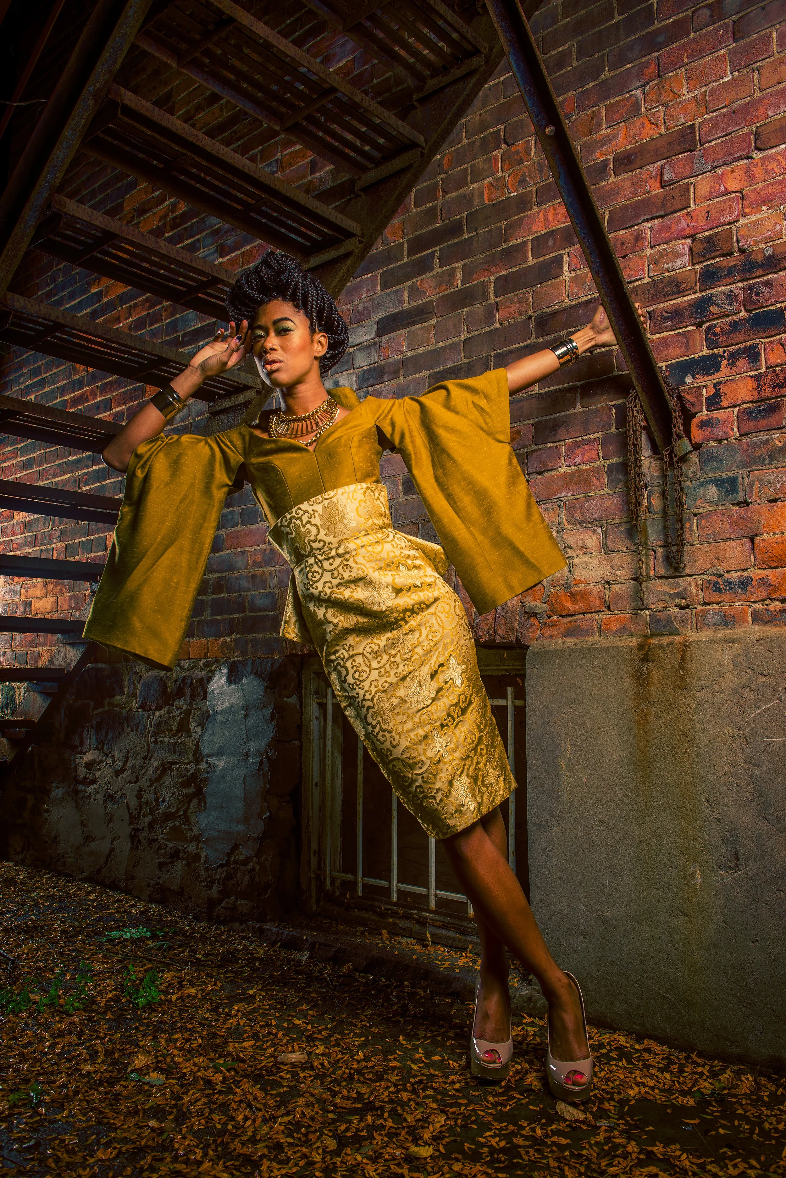 A woman in a gold-patterned dress and a mustard-colored jacket posing in an alley with a brick wall and staircase. She is accessorized with chunky jewelry and wearing high-heeled shoes, standing on a leaf-strewn ground.