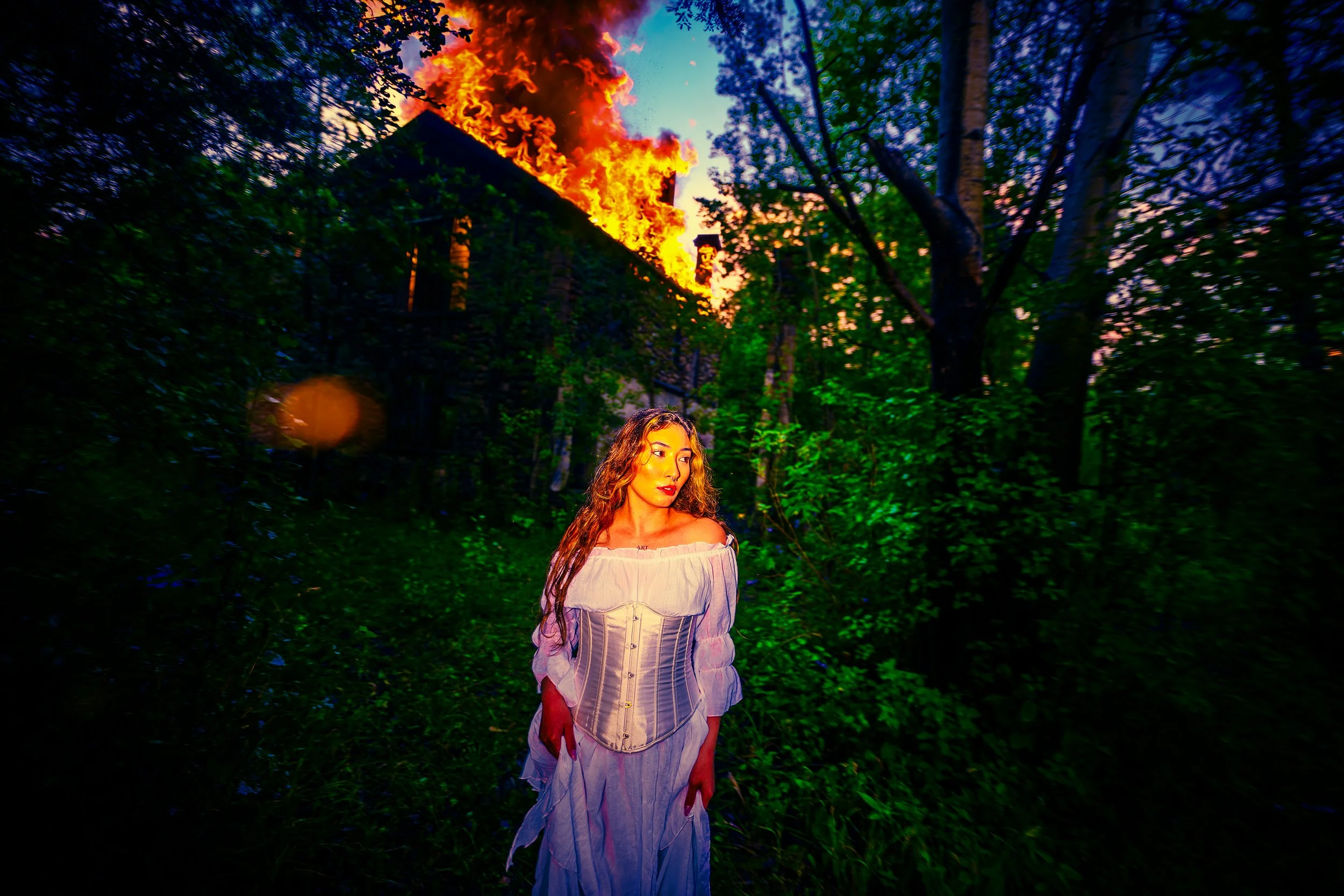 A young woman with wavy hair in a white off-the-shoulder top and corset stands in a wooded area at sunset. Behind her, a house is on fire with large flames and smoke billowing into the sky.