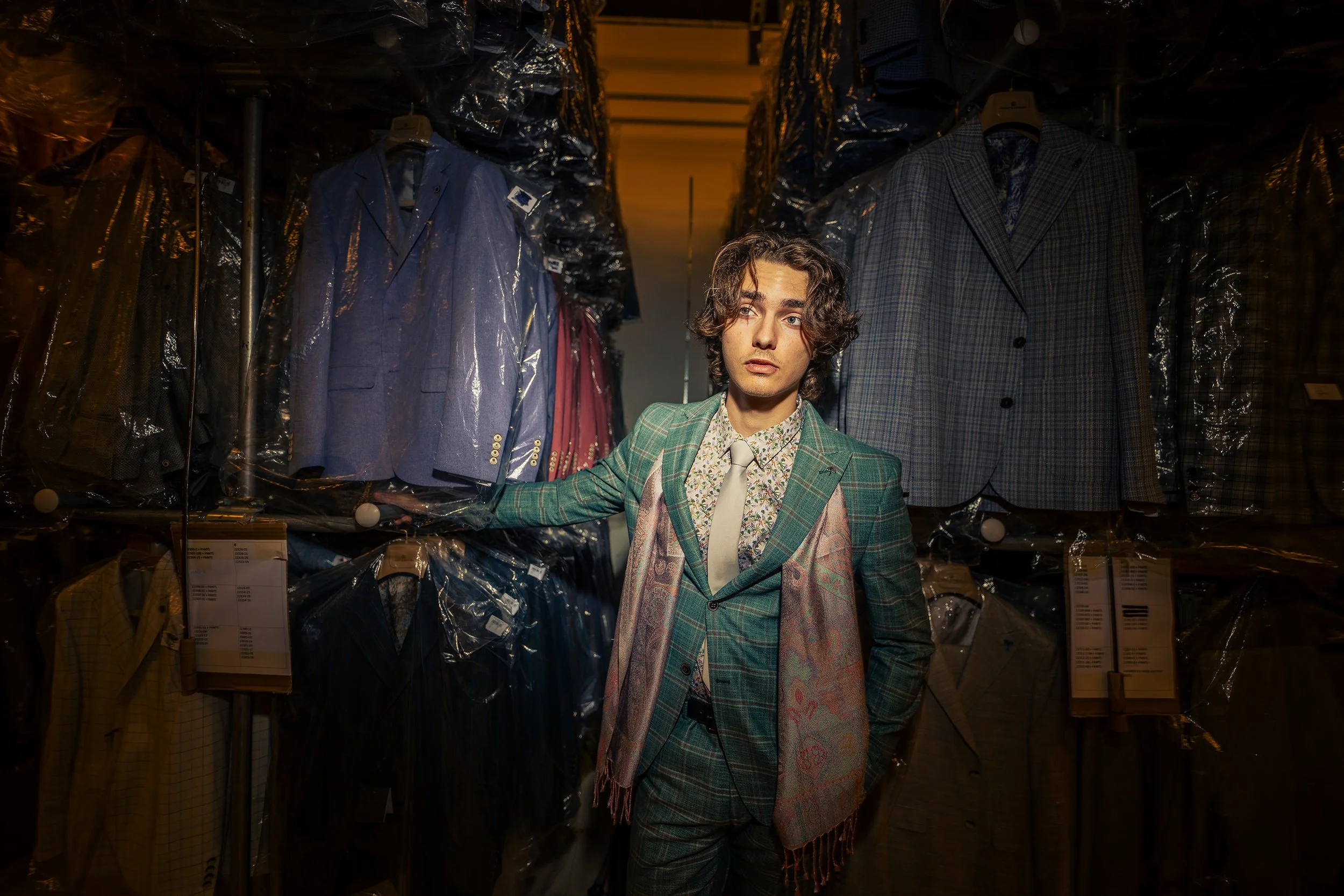 A young man with curly hair and a plaid green blazer posing among racks of suits in a boutique.