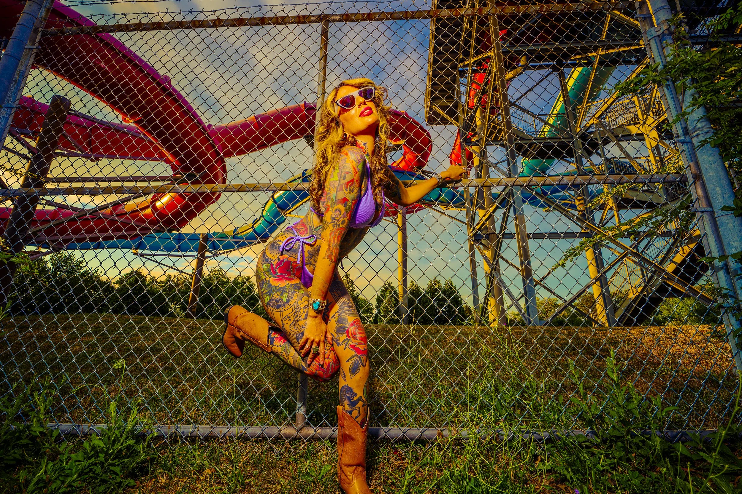 A tattooed woman wearing sunglasses, purple bikini, and brown boots, posing behind a chain-link fence at an amusement park with a large colorful waterslide structure in the background during sunset.