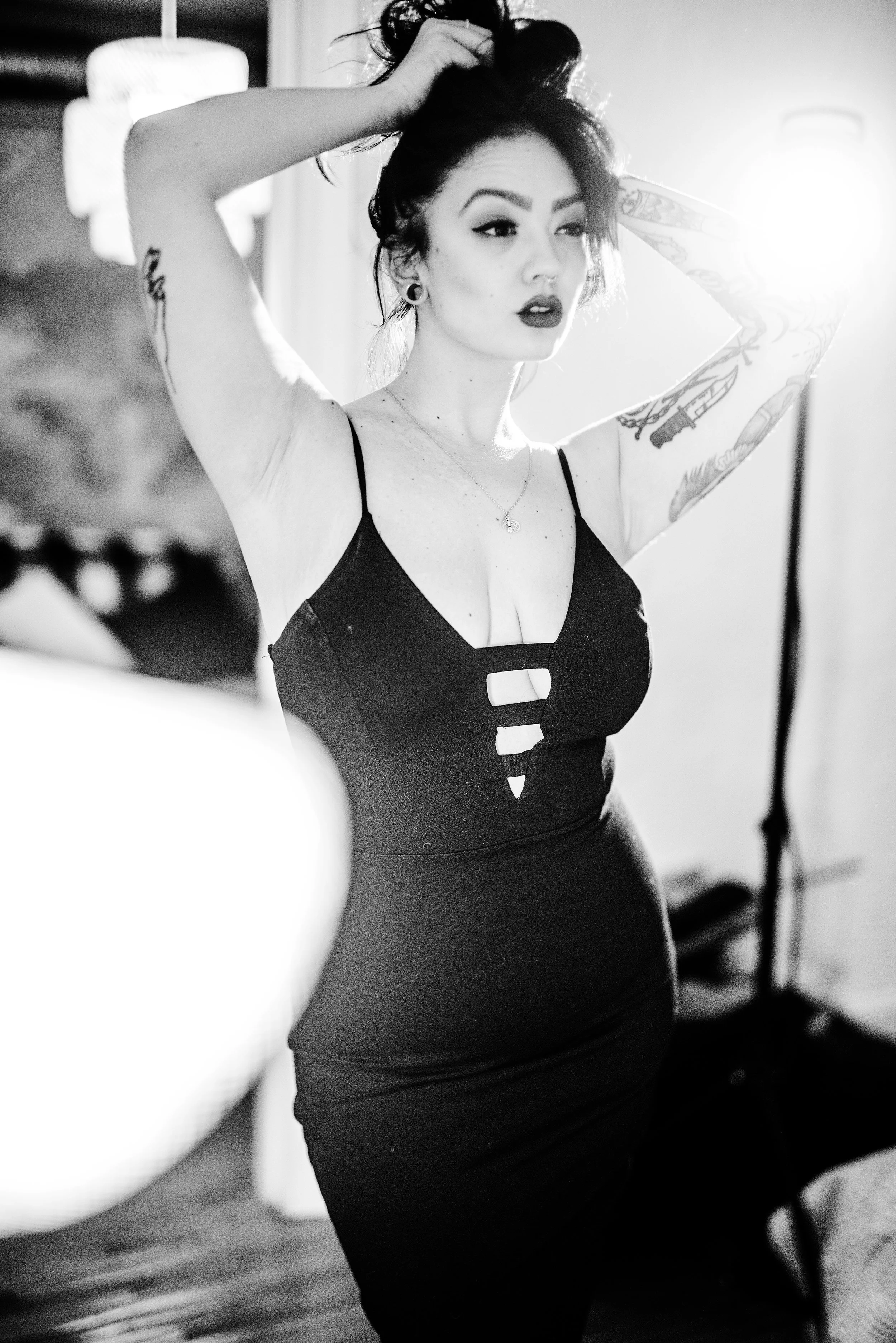 A woman with tattoos wearing a black dress, standing indoors with one hand on her head, looking to the side, with a bright light in the background.