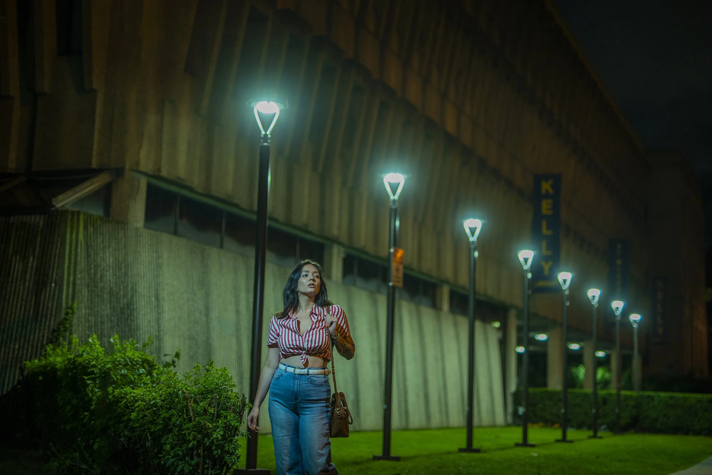 A woman with dark hair stands outside at night under streetlights, wearing a red and white striped shirt tied at the waist and blue jeans, holding a brown purse and looking to the side near a building with a sign that reads 'KELLY'.
