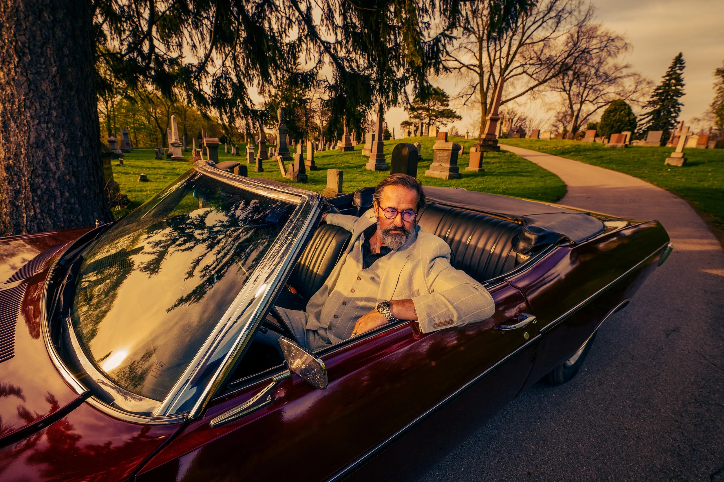 A man with glasses and a beard in a beige suit takes a selfie while sitting in a vintage red car in a cemetery at sunset, with headstones and trees in the background.