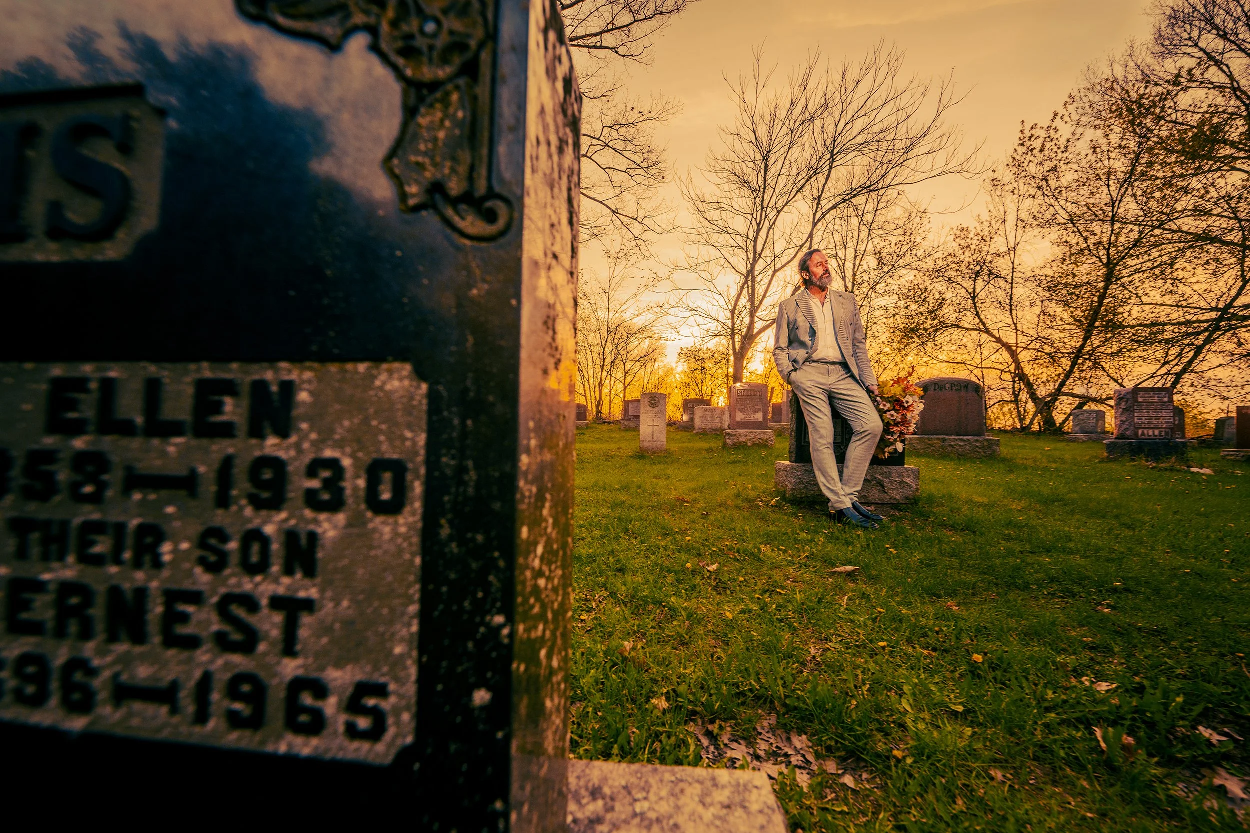 A man in a gray suit standing in a cemetery at sunset, with leafless trees and gravestones in the background. A close-up of a cemetery marker in the foreground.