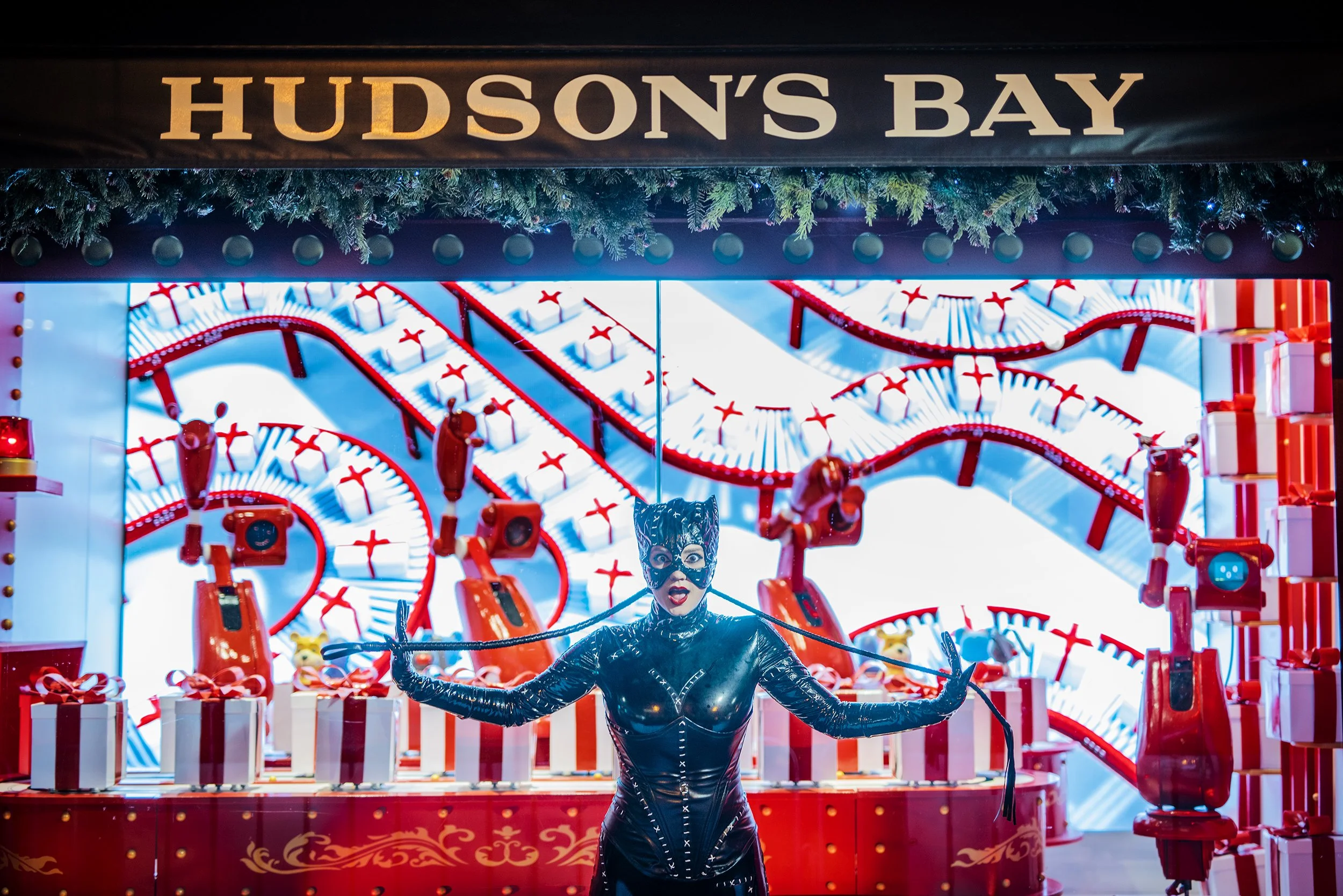 A person dressed in a black latex catsuit and wearing a mask stands on a festive display with a sign that reads 'Hudson's Bay.' The display features a large roller coaster model decorated with Christmas ornaments, gift boxes, and greenery.