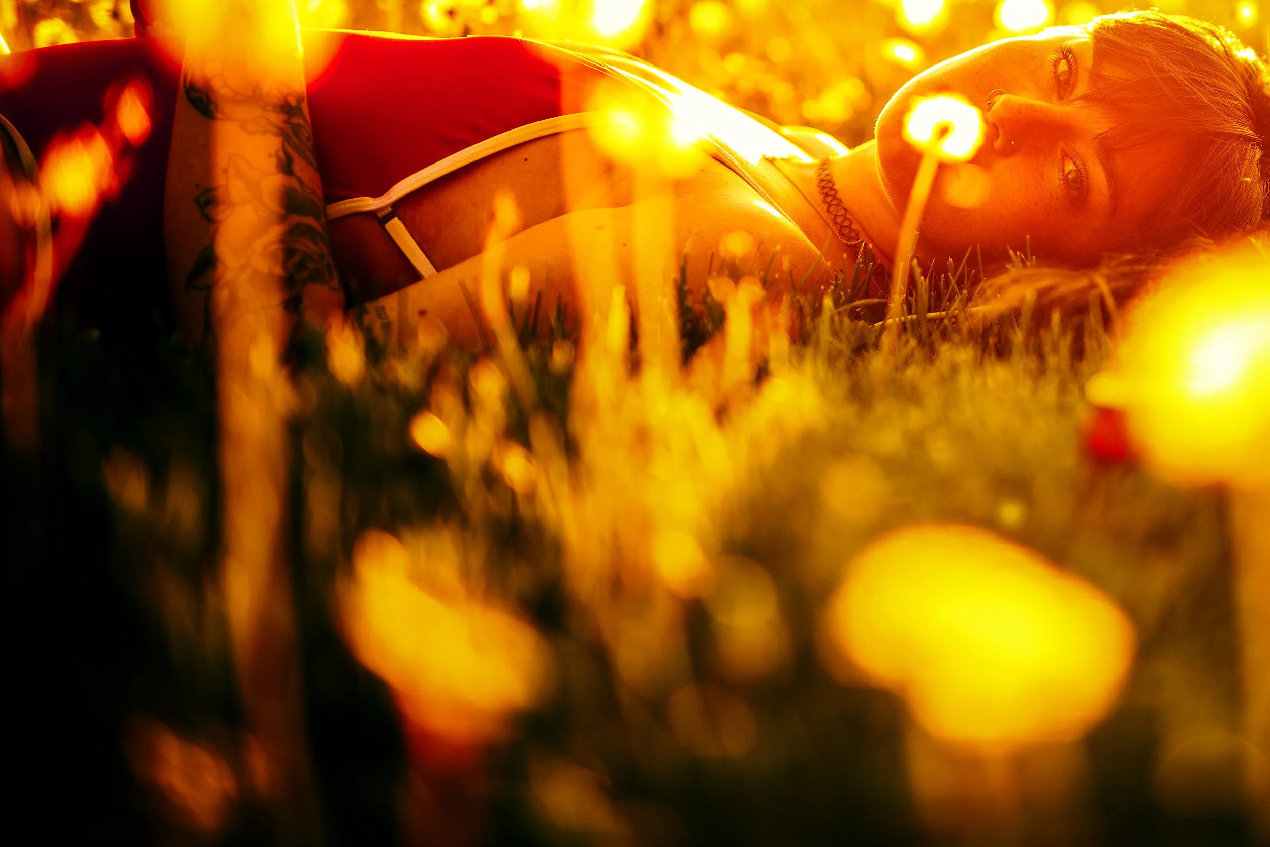 A woman lying on the grass during sunset, with warm orange and yellow tones, looking at the camera, wearing a red top, with tattoos on her arm and a choker necklace.