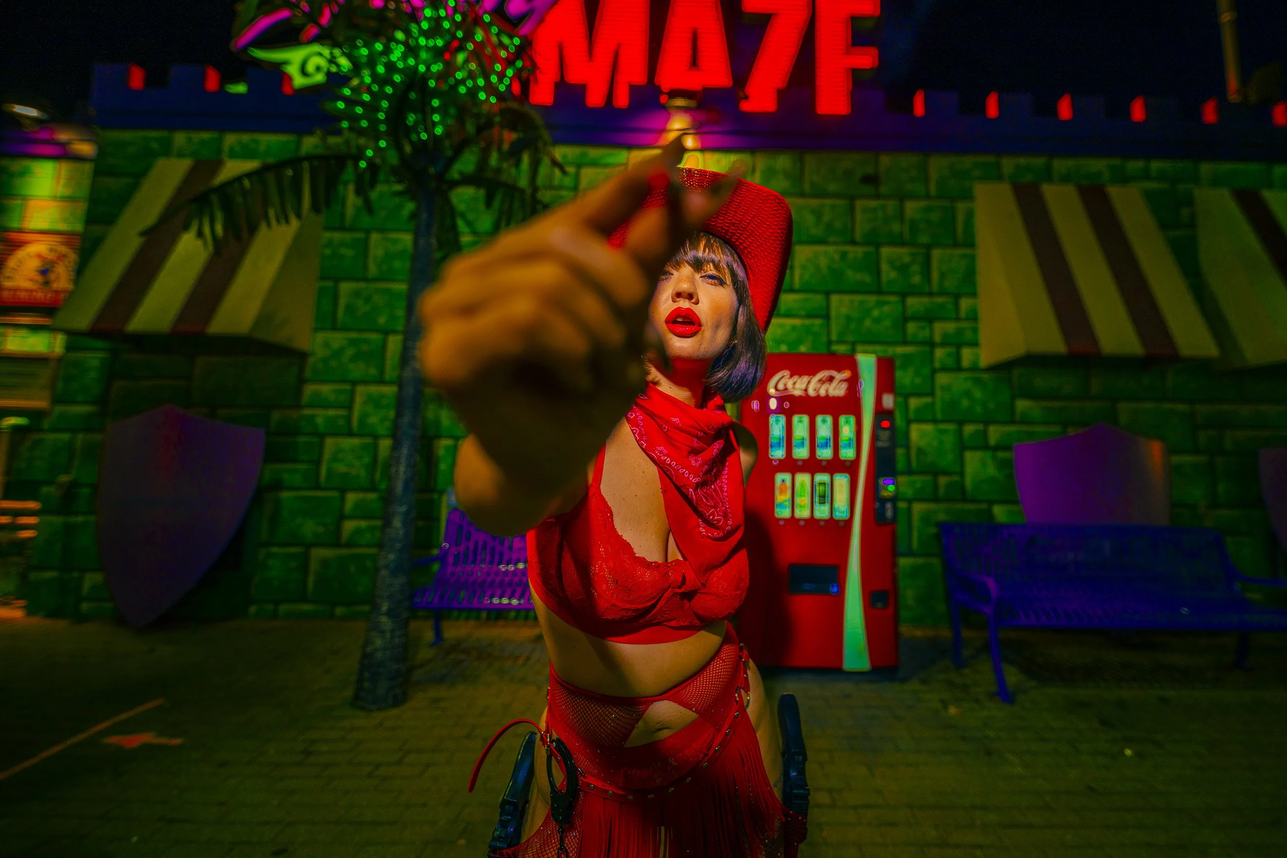 A woman dressed in red, wearing lingerie with fishnet stockings and a red bandana, pointing towards the camera in front of a green brick wall, with colorful neon signs and a Coca-Cola vending machine in the background.