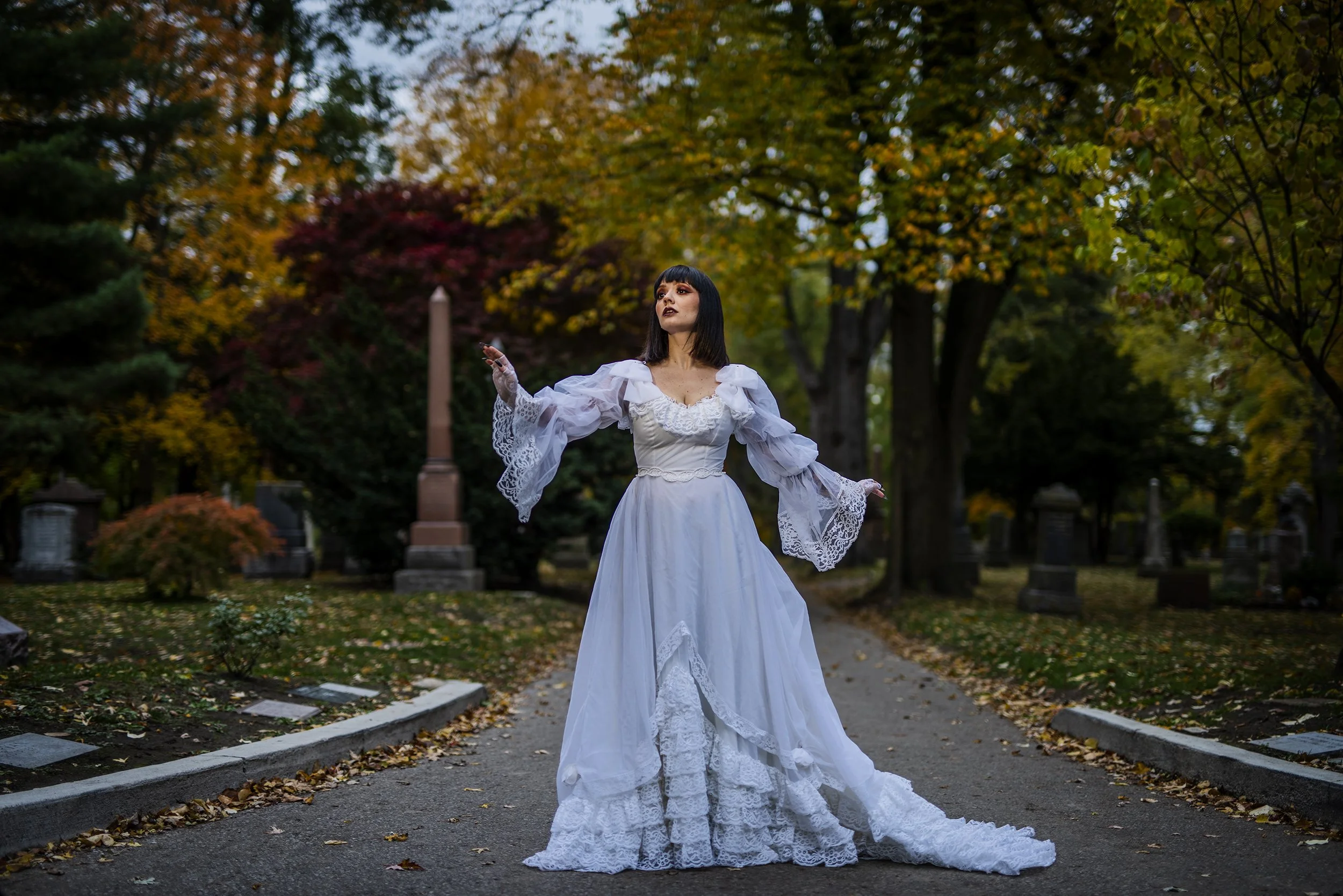 A woman in a vintage white wedding dress with lace details stands in a cemetery during fall, surrounded by colorful trees with yellow and red leaves. She has dark hair and makeup, and is holding a small object in her right hand, with a serious expres