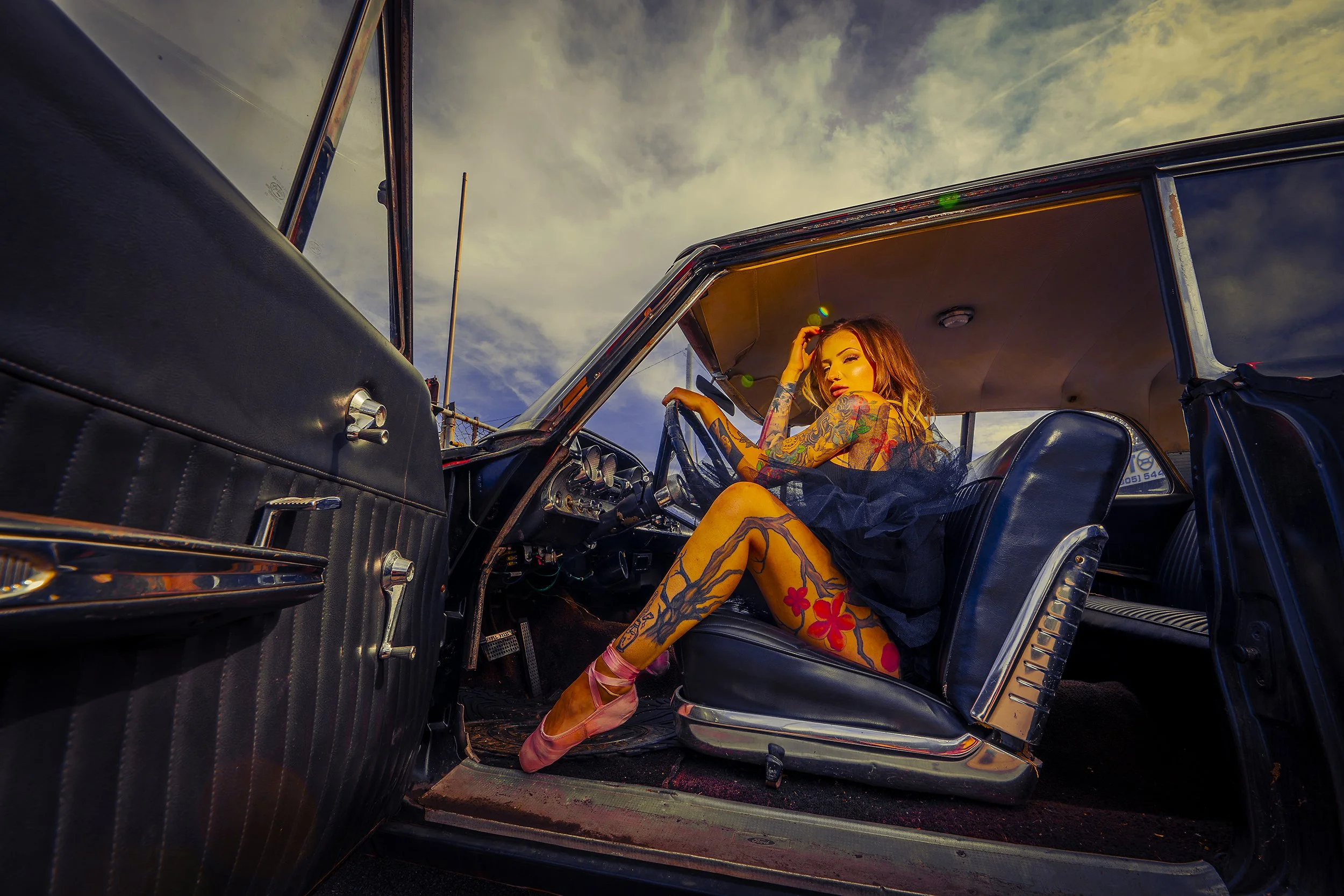 A woman with tattoos sitting on the front seat of a vintage car, wearing a black dress and pink ballet shoes, touching her hair with one hand, under a cloudy sky.
