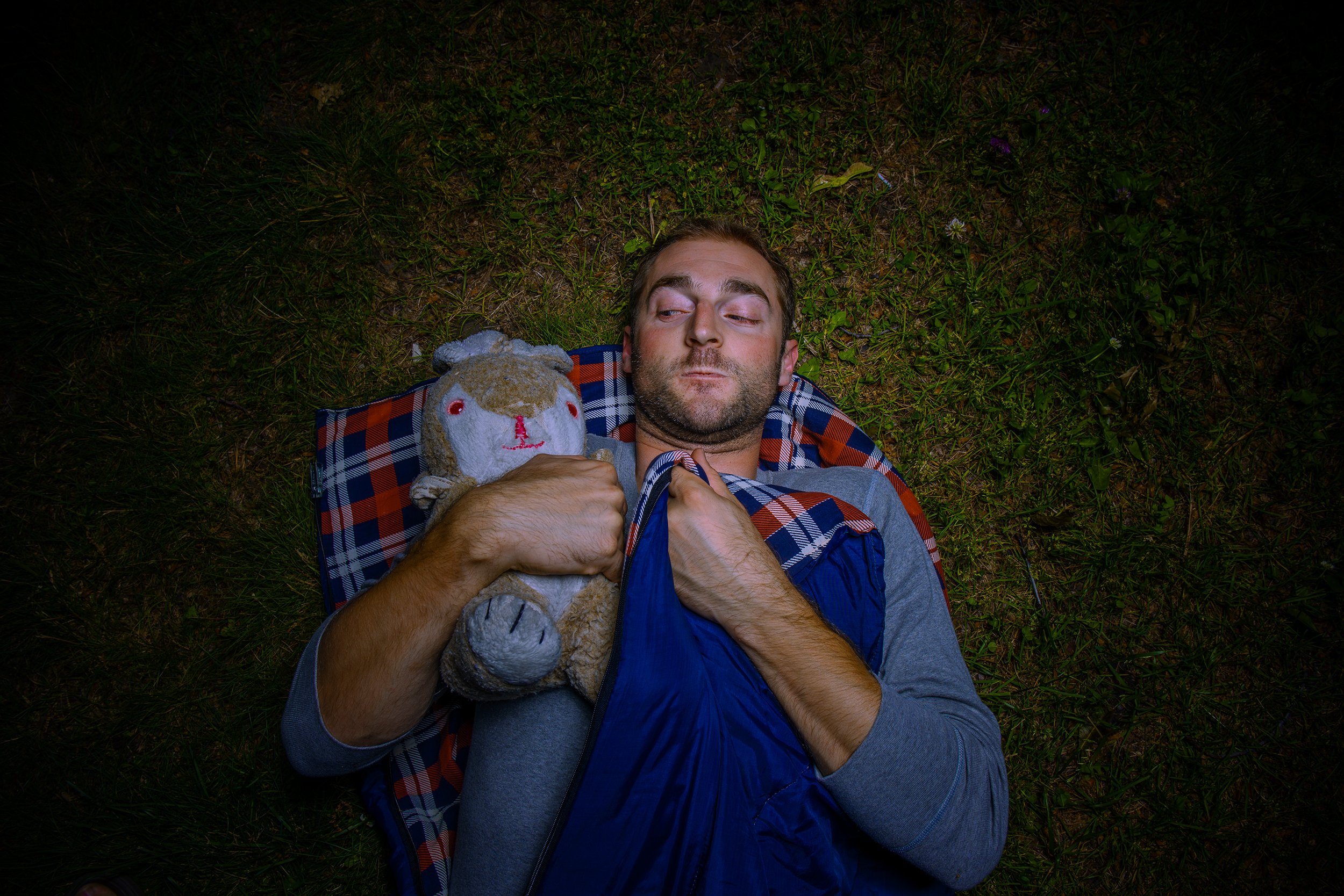 A man lying on the grass, holding a stuffed animal bear and a blue bag, with a plaid shirt and a gray sweater, looking to the side.