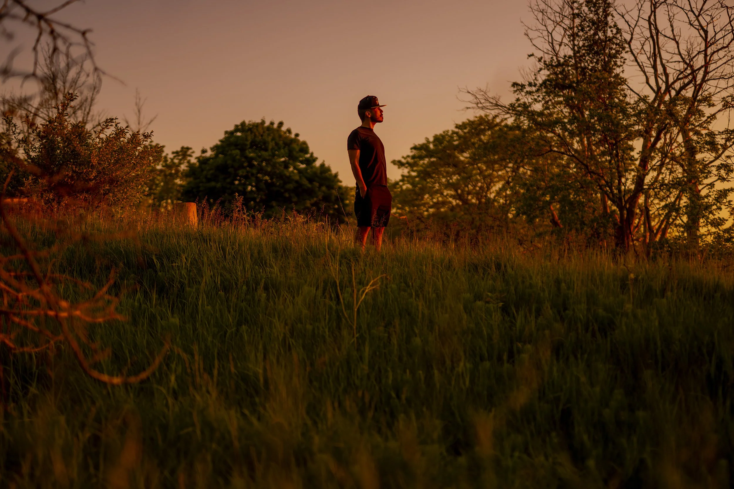 A man standing in a grassy field at sunset with trees in the background.