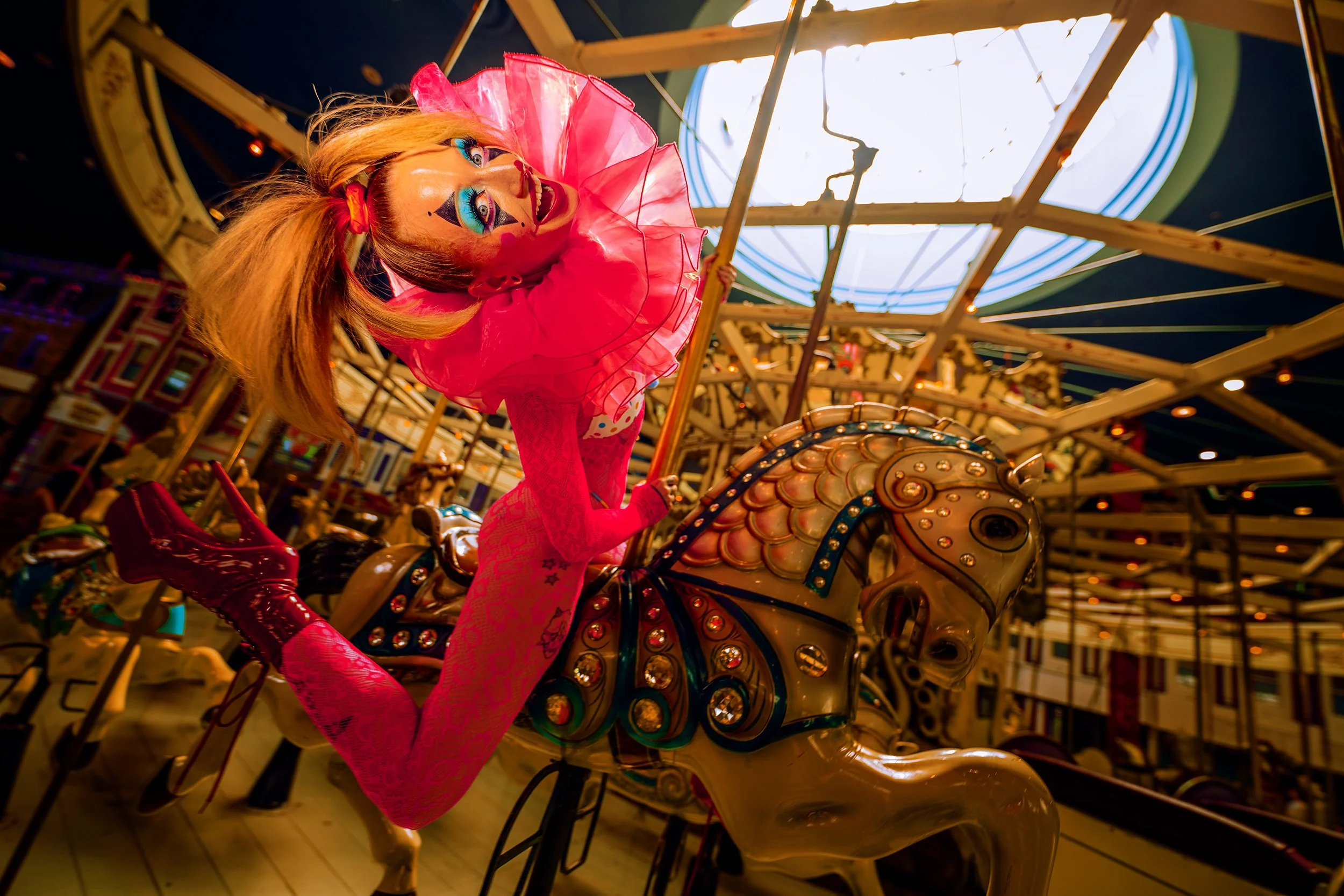 A performer with bright clown makeup riding a carousel horse in a carnival setting, wearing pink clothing and a pink ruffled collar.