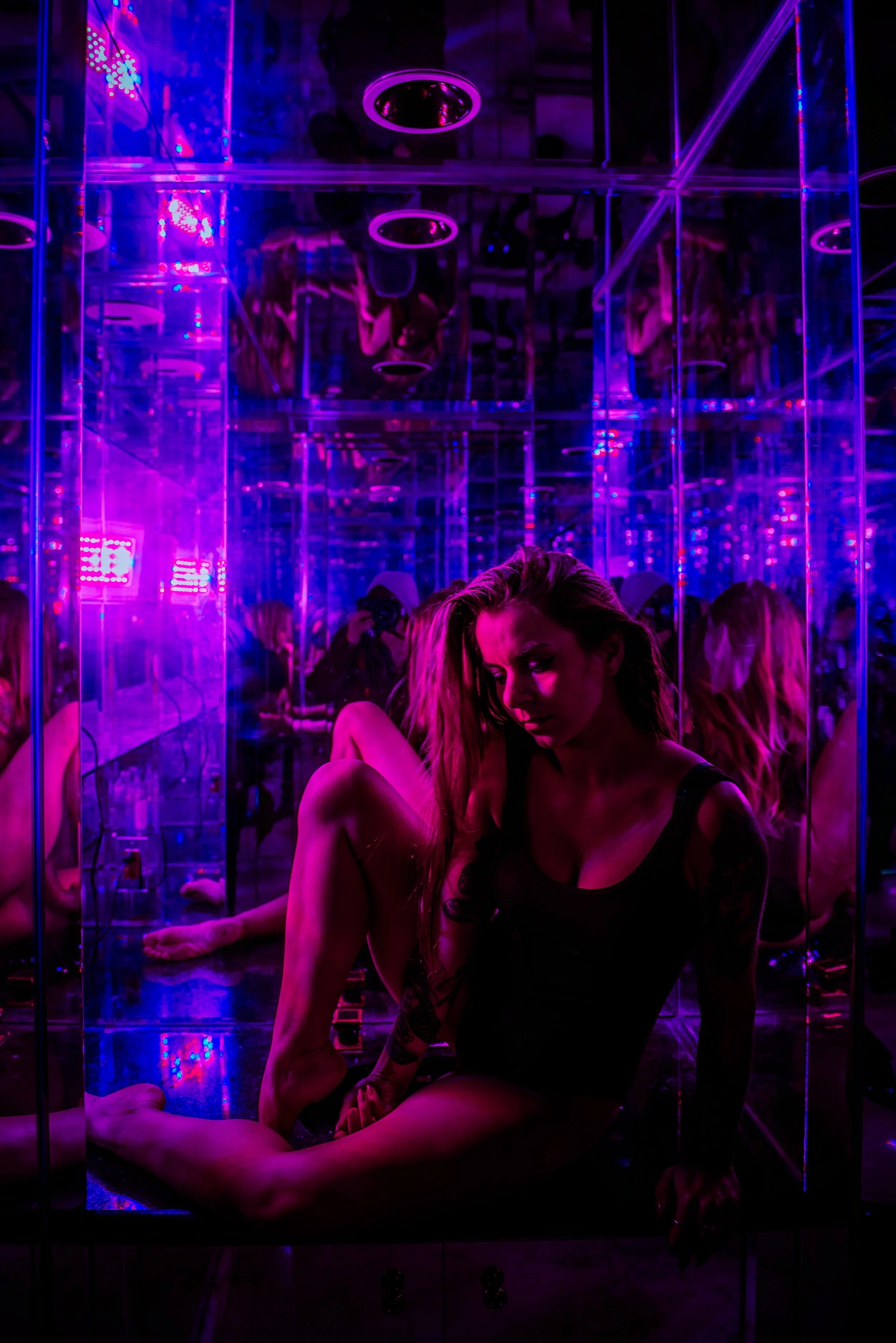 A woman with long hair and tattoos sitting on the floor of a mirror-lined room illuminated with purple and pink lights, with reflections of herself and others visible in the mirrors.