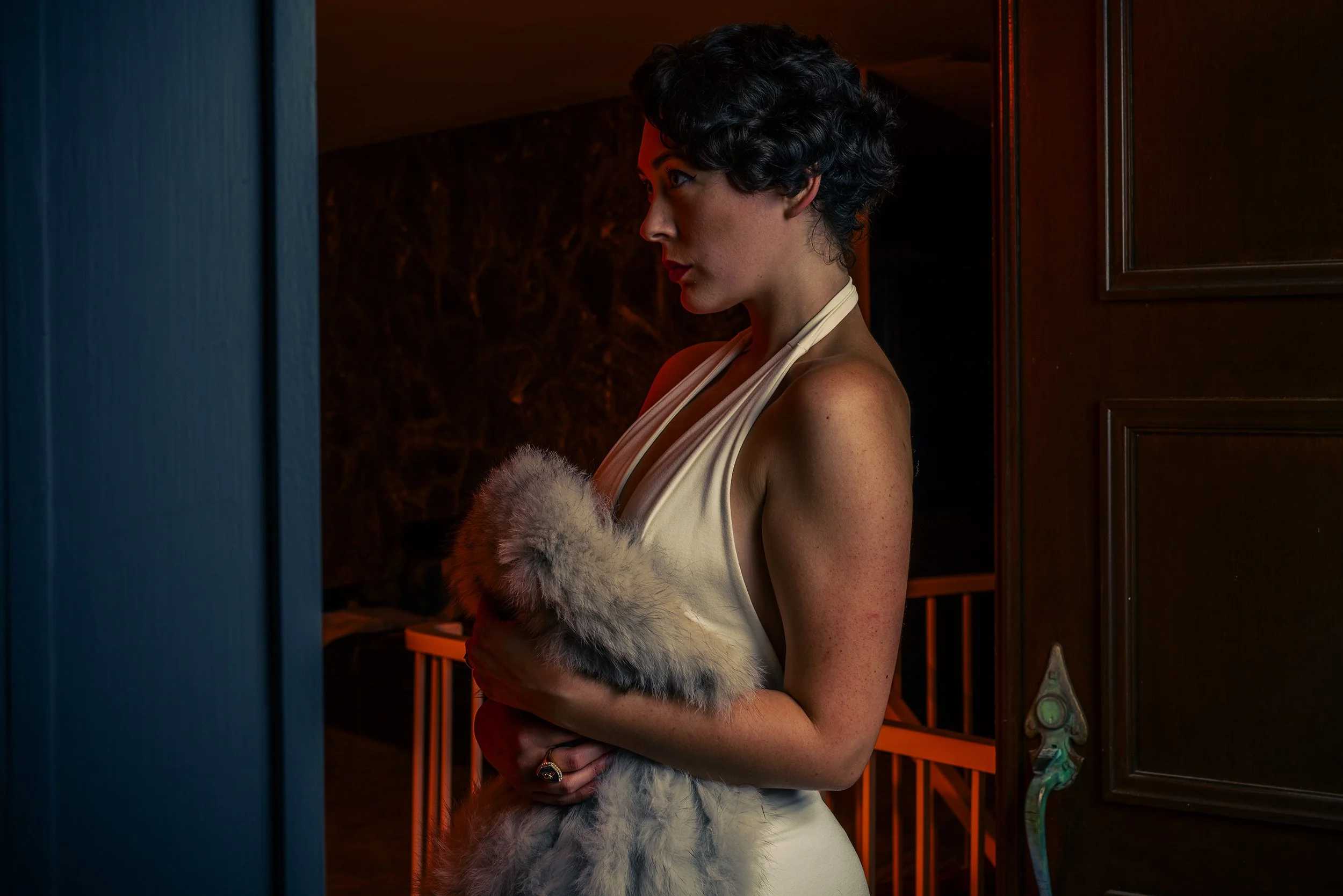 Woman with short curly dark hair wearing a white dress and a fur stole, standing in a dimly lit room, looking to her left.