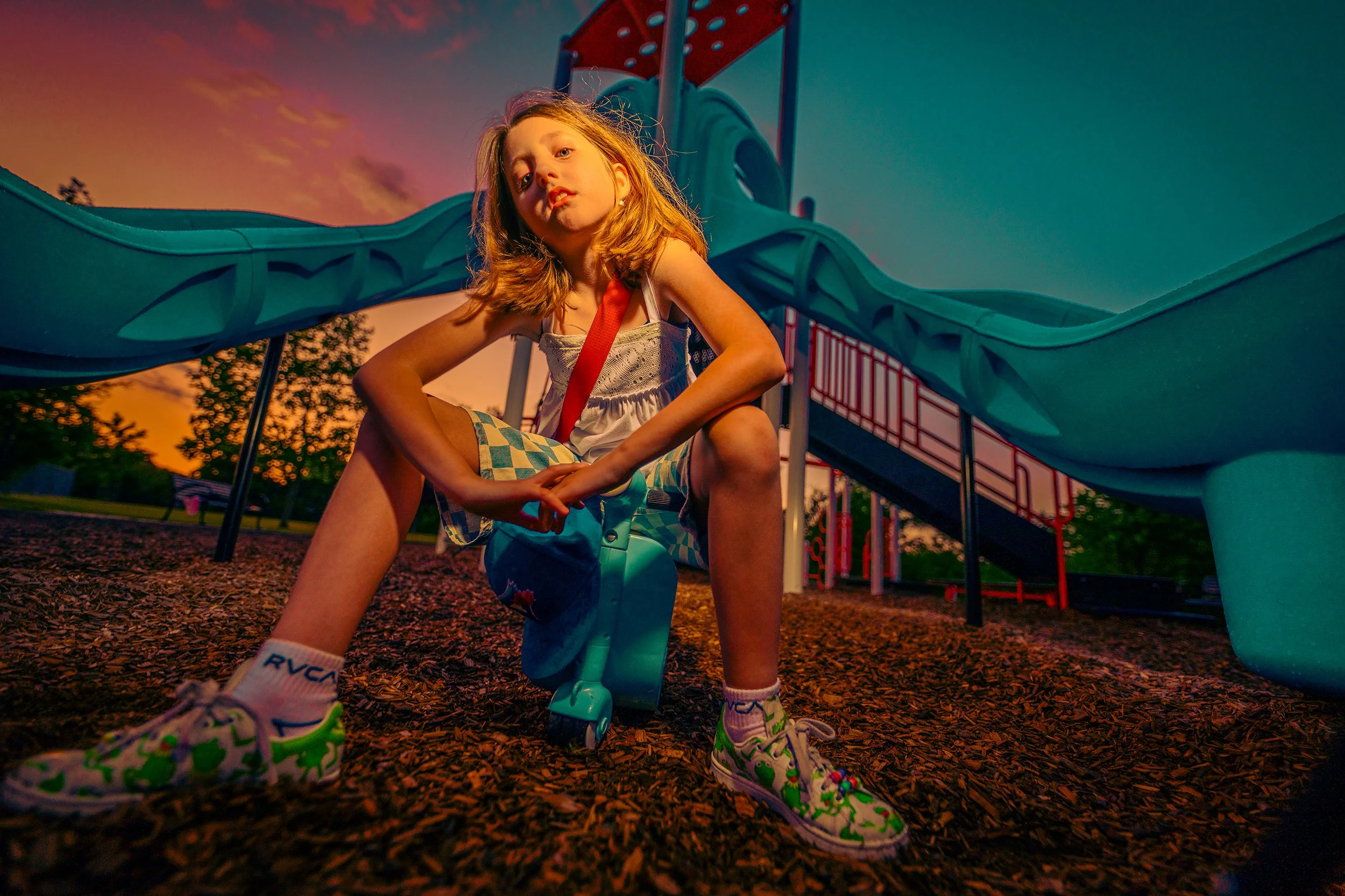 A young girl with blonde hair sits on the ground at a playground, with a slide in the background during sunset. She is wearing a white tank top, checkered shorts, and colorful sneakers. The shot is taken from a low angle, emphasizing her in the frame