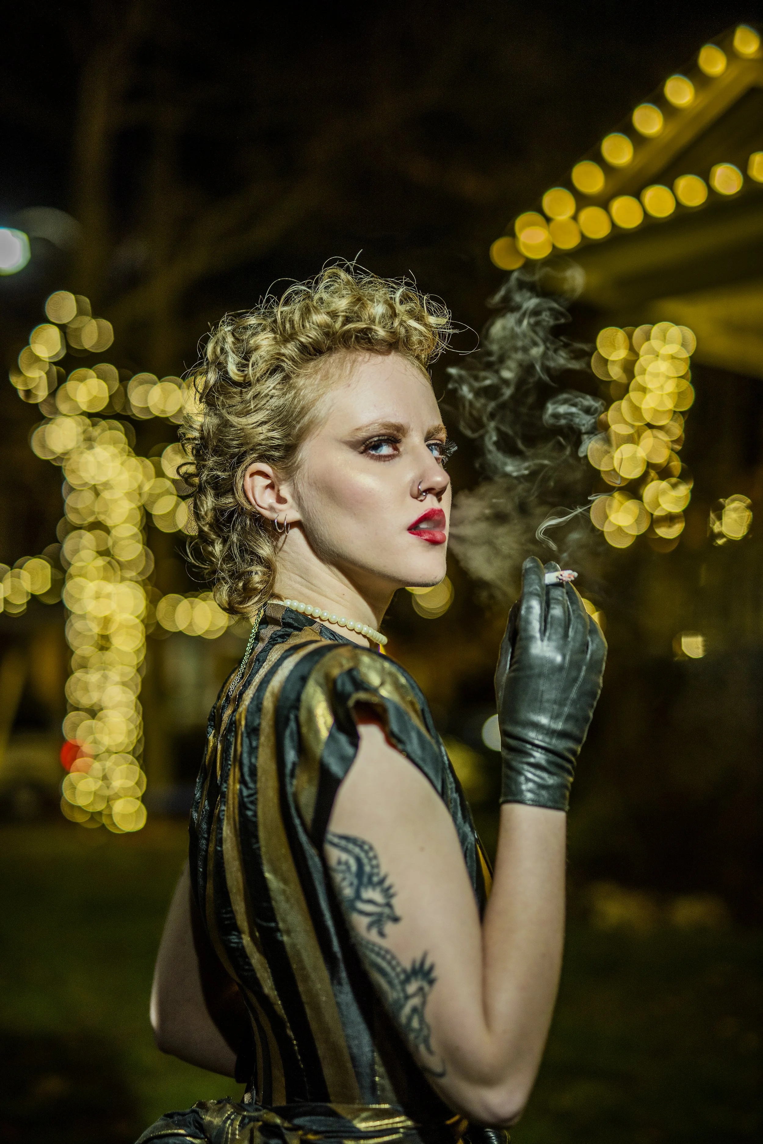 Young woman with curly blonde hair, blue eyes, red lipstick, and tattoos on her arm, smoking a cigarette at night with blurred yellow lights in the background.