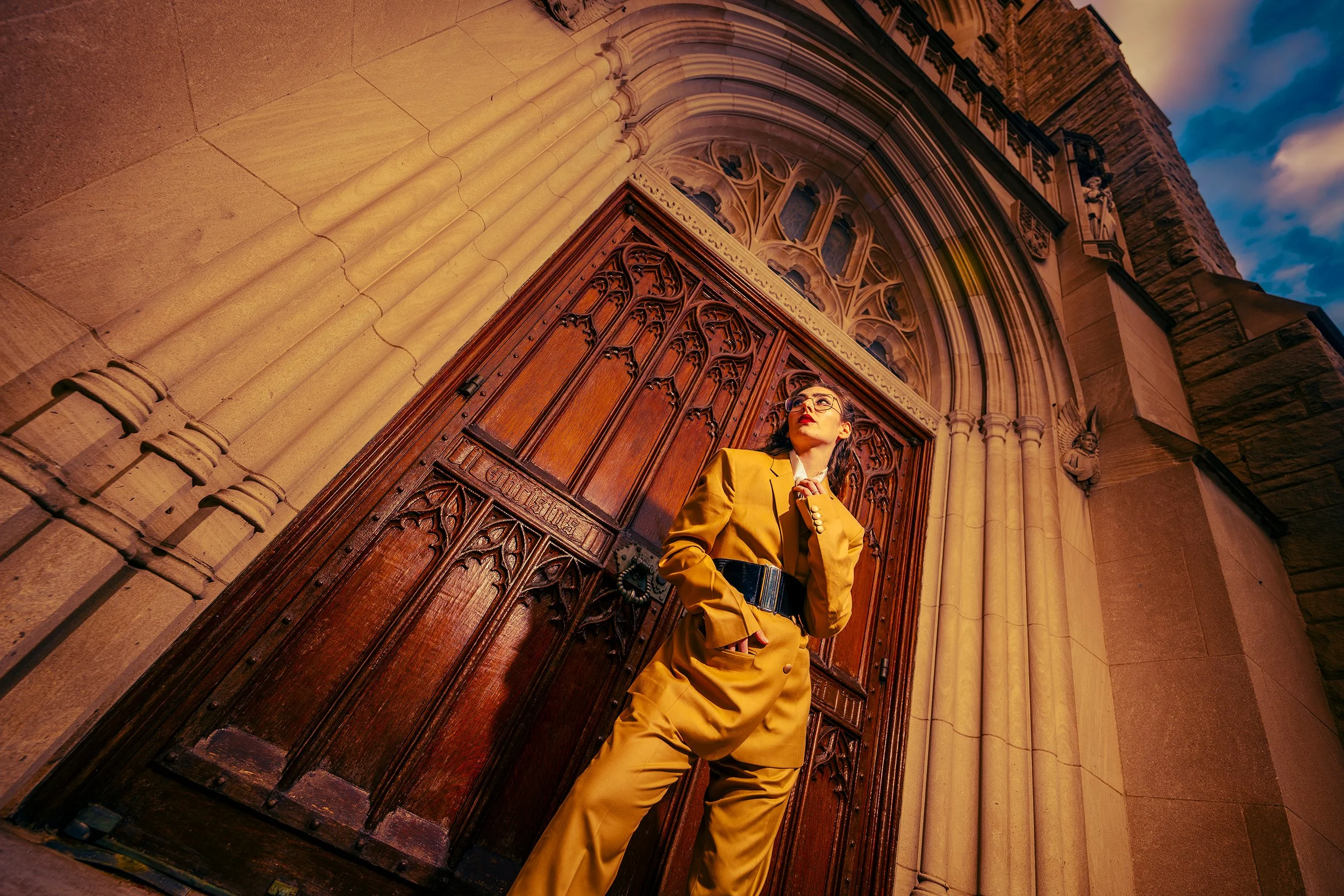 A woman in a yellow suit standing in front of a Gothic church entrance with ornate wooden doors and stone columns, looking up at the sky during sunset.