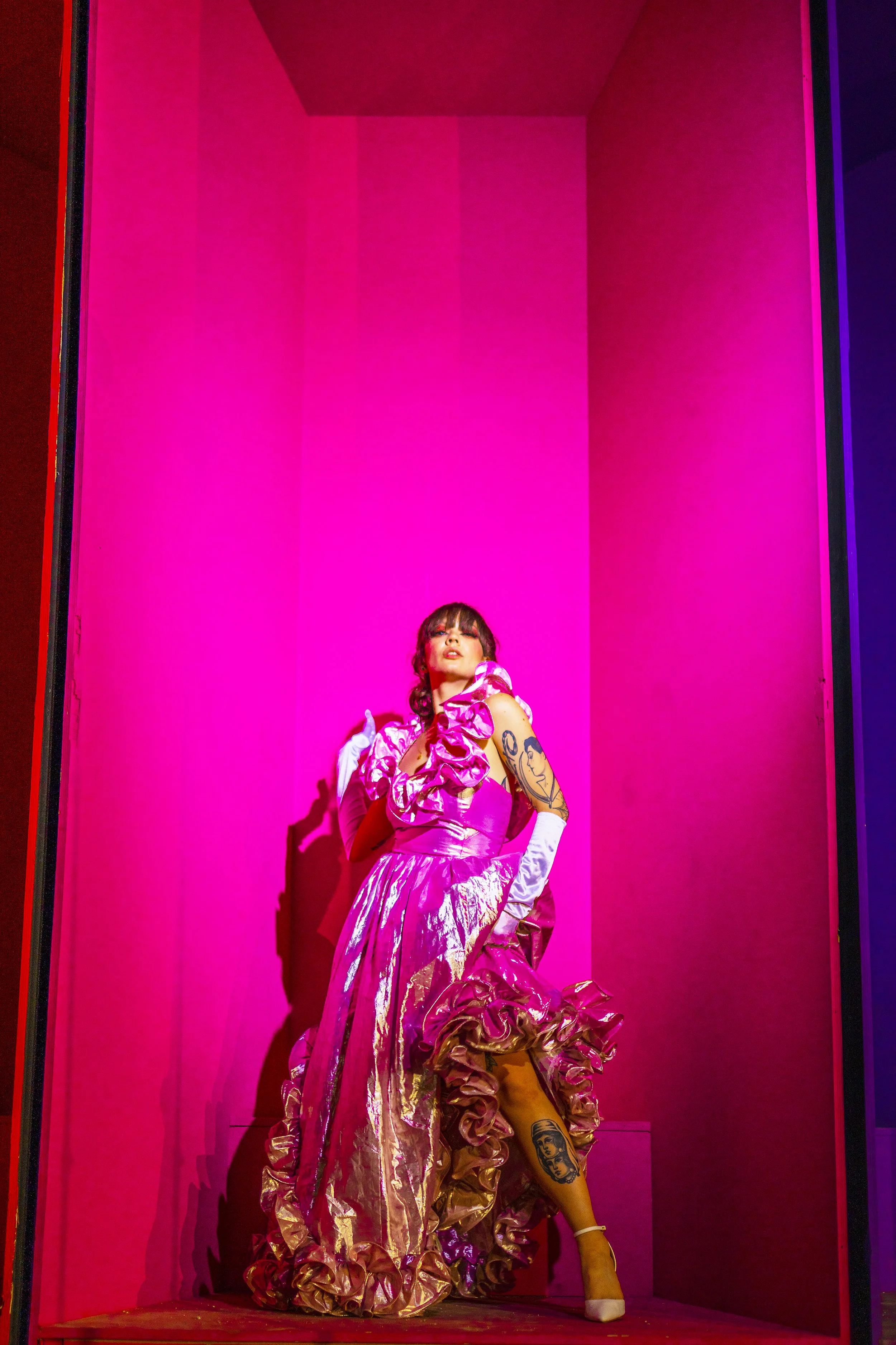 A woman dressed in a pink, ruffled gown stands in a small pink-lit display case, with tattoos on her arm and leg, wearing white gloves and beige heels.