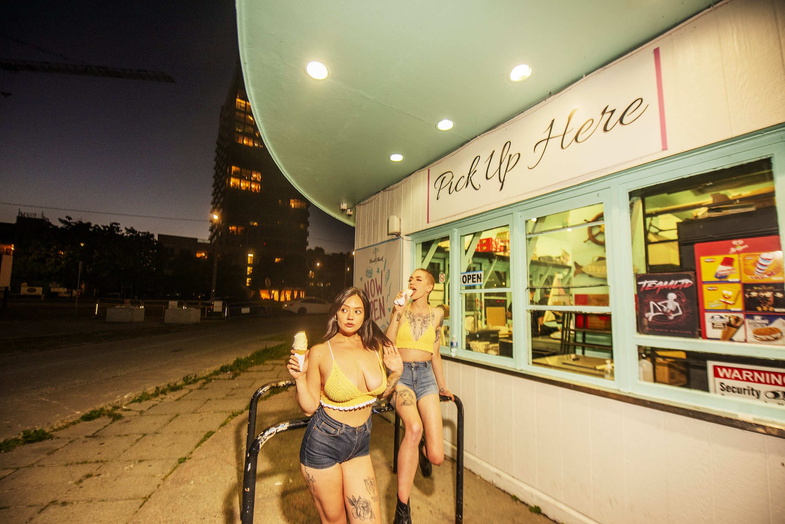 Two women in yellow tops and denim shorts standing outside an ice cream shop at night, one holding ice cream and the other eating ice cream.
