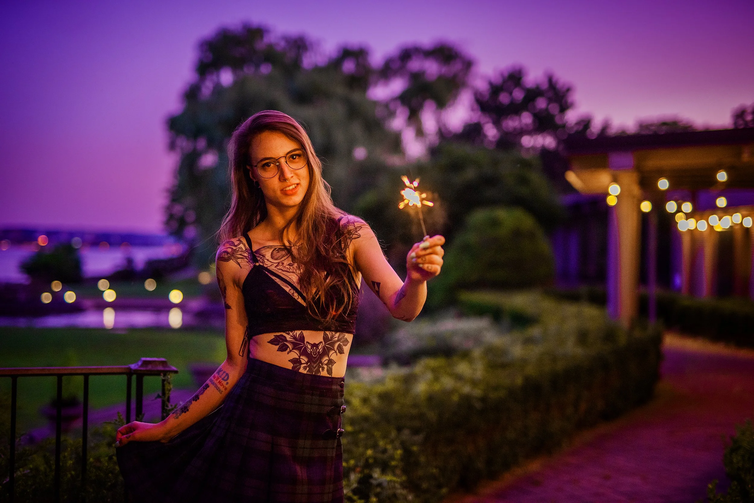 A young woman with long hair, glasses, and tattoos wearing a black lace crop top and plaid skirt, holding a lit sparkler in an outdoor setting during dusk or evening with purple sky and blurred background of trees and lights.