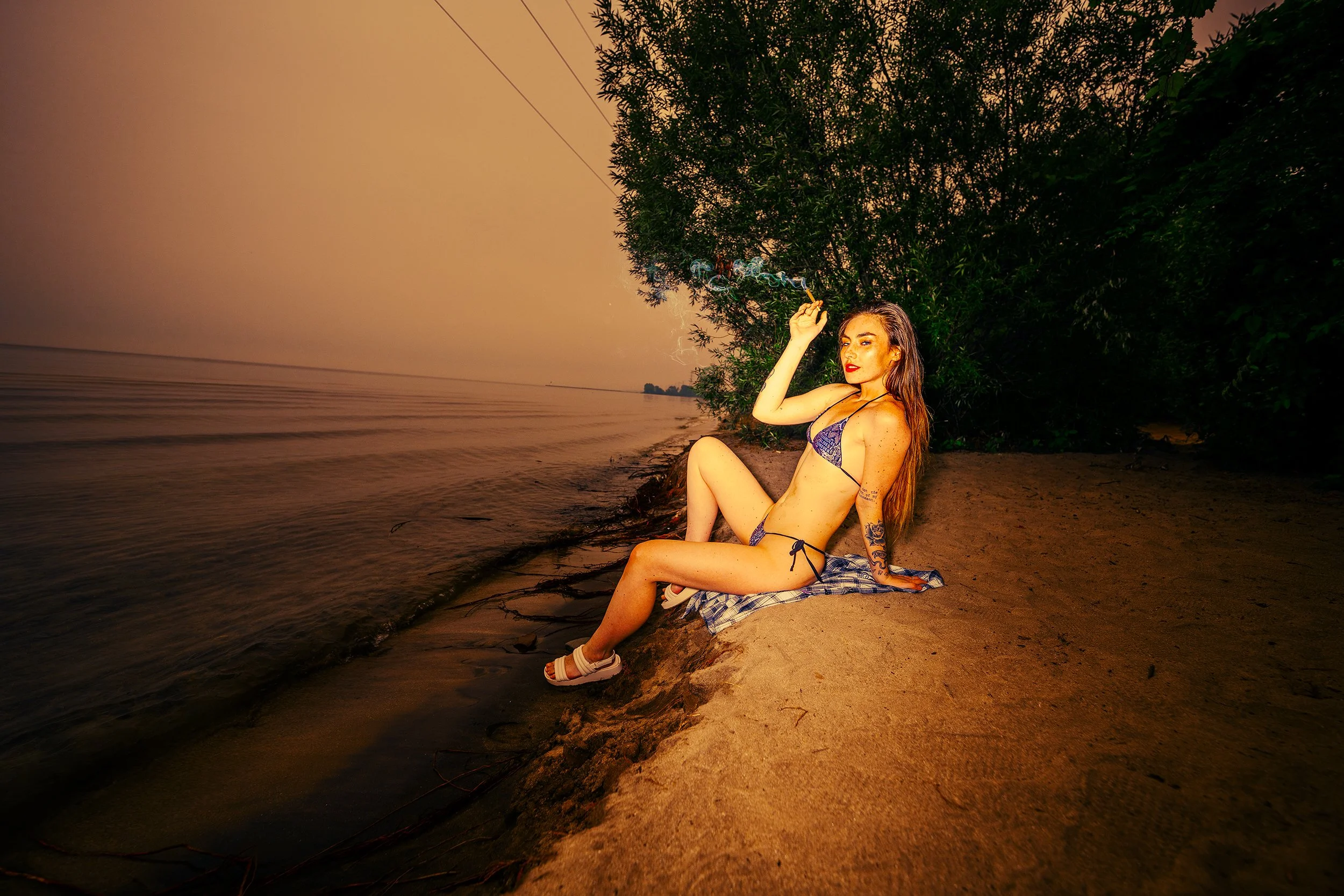 A woman sitting on a sandy beach, wearing a bikini, smoking a cigarette, and looking at the camera as she leans against her hand. There are trees in the background and the sun appears to be setting, creating an orange hue in the sky and reflecting on