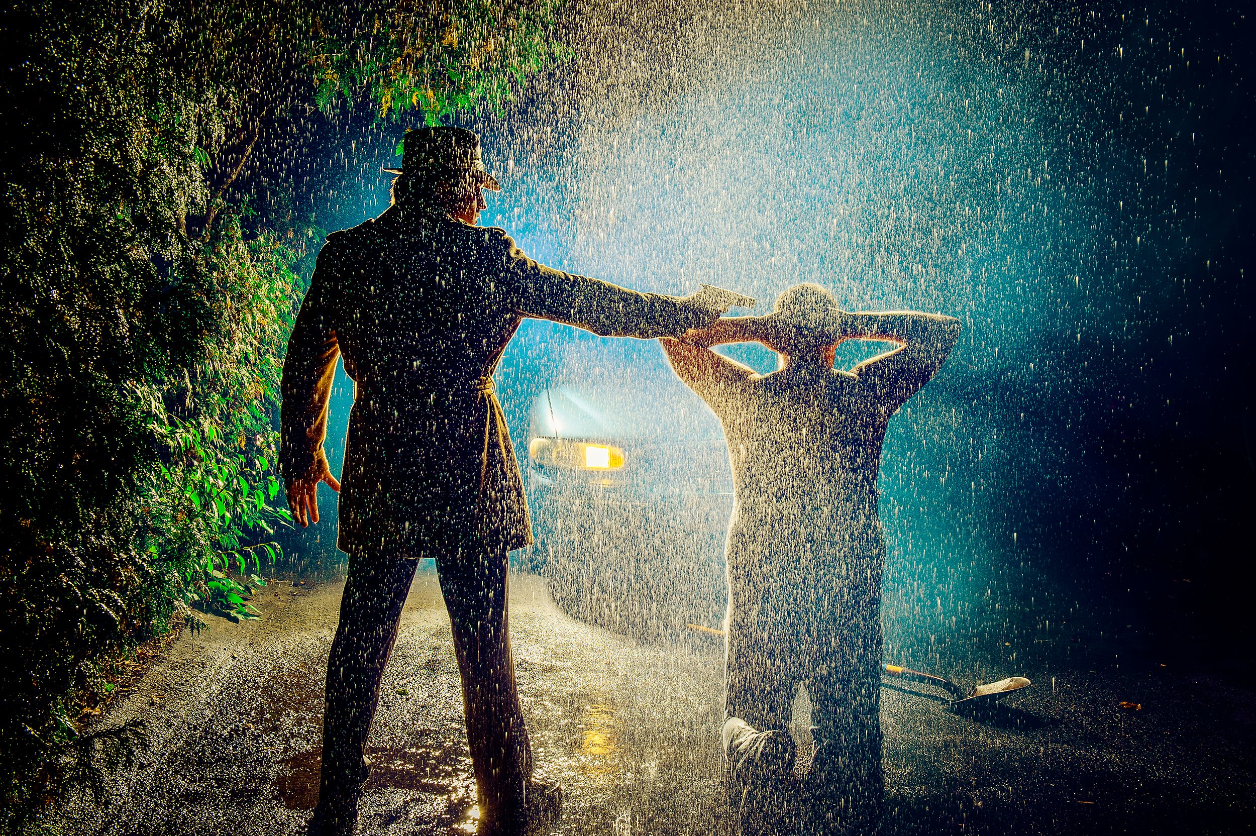 Two children and an adult playing with water under rain at night, illuminated by streetlight, with greenery on the side and a car in the background.