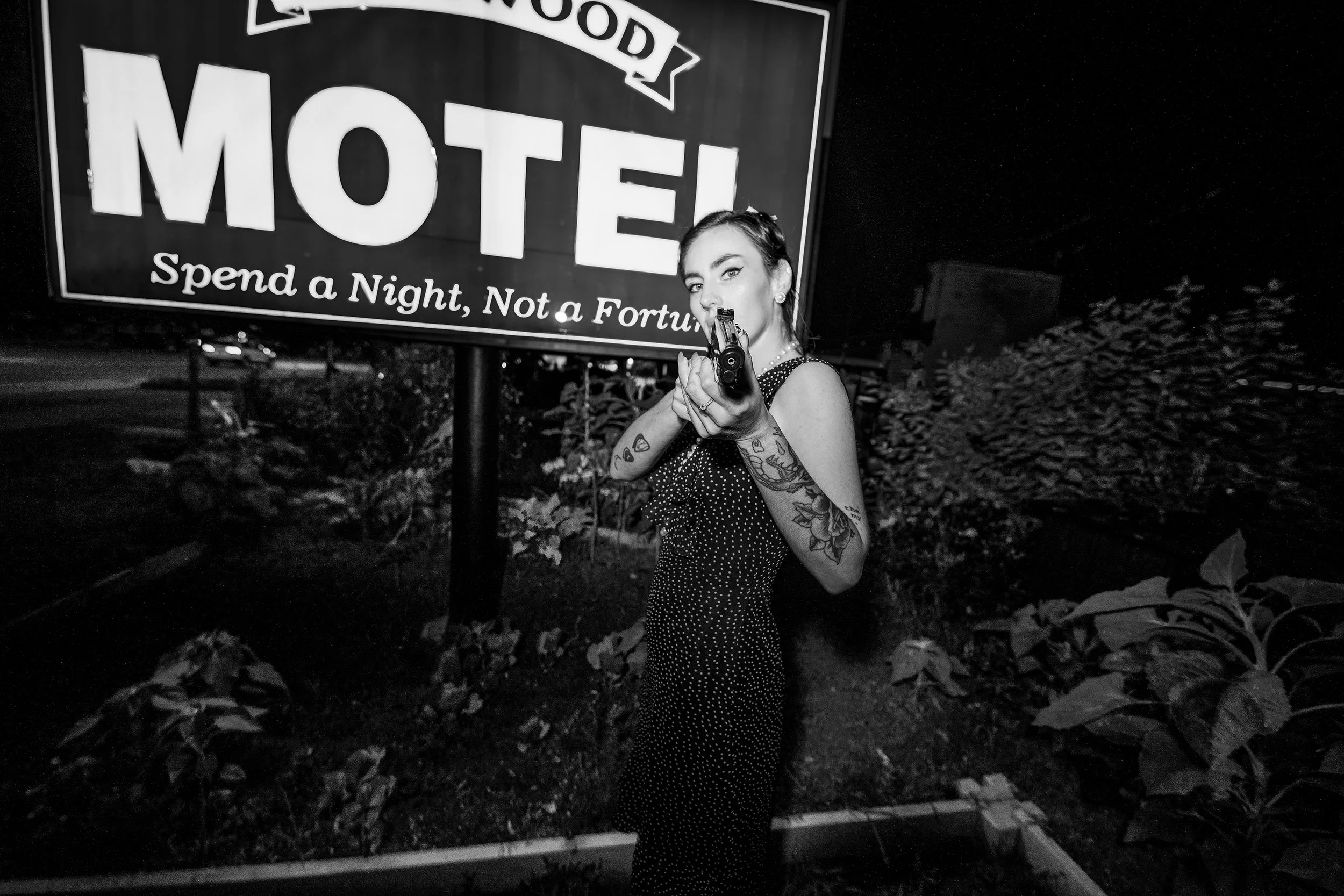 A woman with tattoos on her arm holding a gun, standing in front of a motel sign at night.