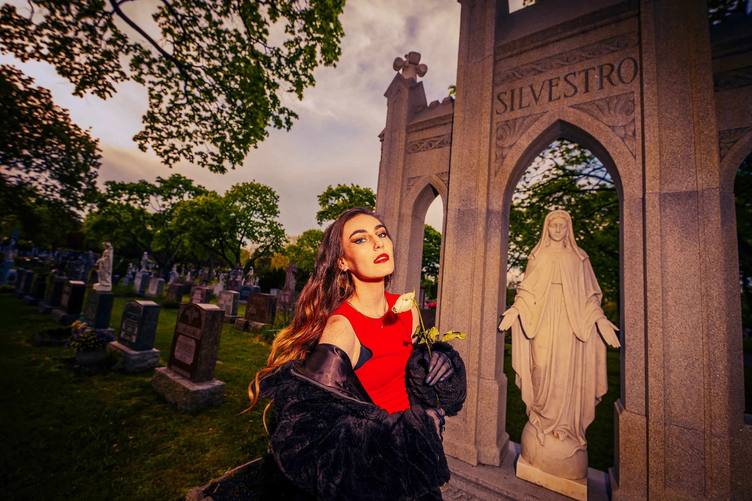 A woman with long wavy hair, wearing a red dress and black gloves, standing in a cemetery at sunset, holding a white rose, next to a religious statue and an arched stone monument with the word 'Silvestro' engraved on it.