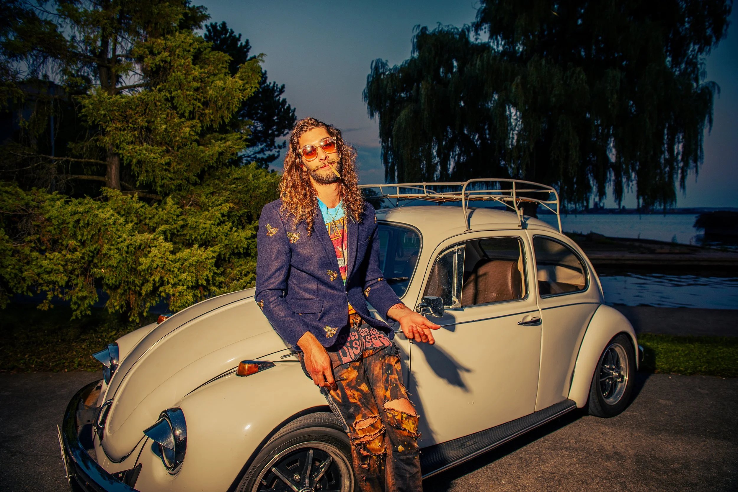 A man with long curly hair, sunglasses, and smoking a cigarette, standing beside a cream-colored vintage Volkswagen Beetle in an outdoor setting during dusk, with trees and water in the background.