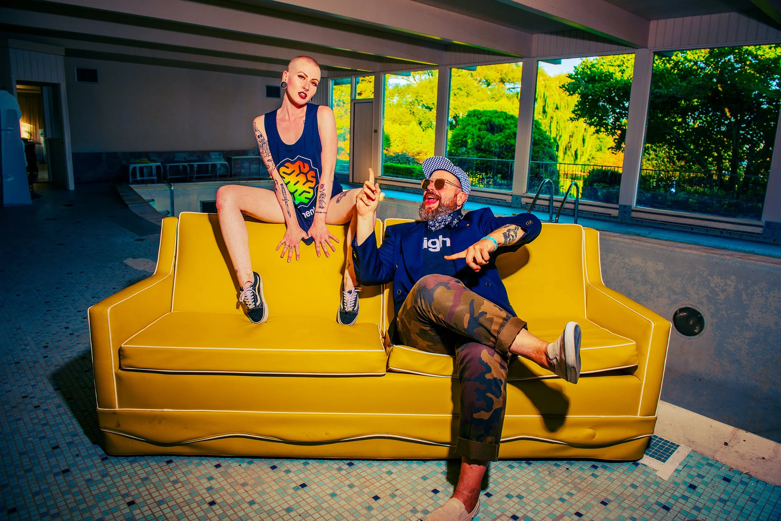 A woman with tattoos and a shaved head sitting on a yellow couch with a man wearing sunglasses, a blue cap, and camouflage pants, in a room with large windows showing trees outside.