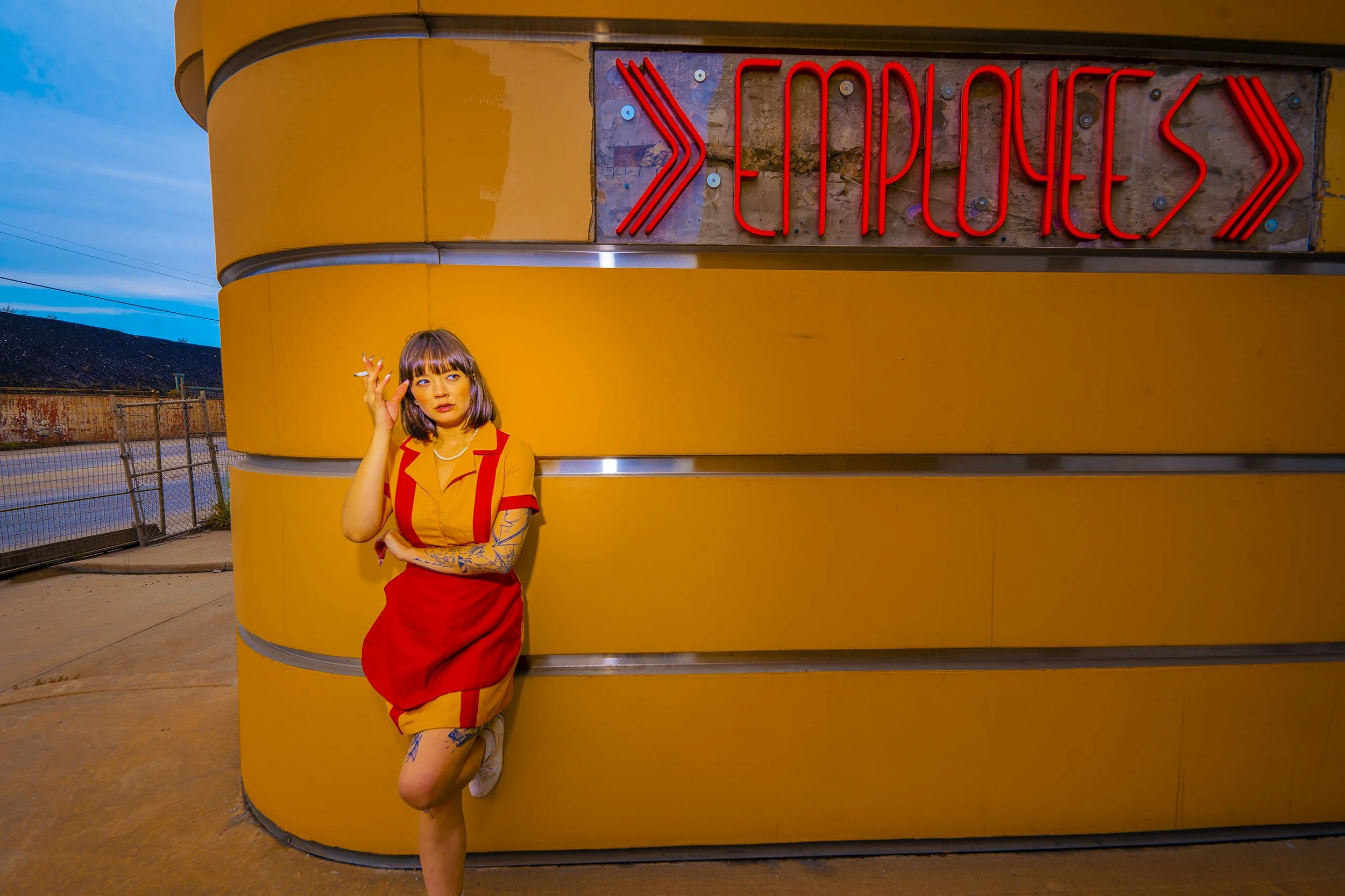 A woman with short dark hair and tattoos on her left arm, wearing a red and yellow dress, stands leaning against a yellow wall near a neon sign that reads 'CAMPUS', holding a cigarette in her right hand. The background features a partly cloudy sky an