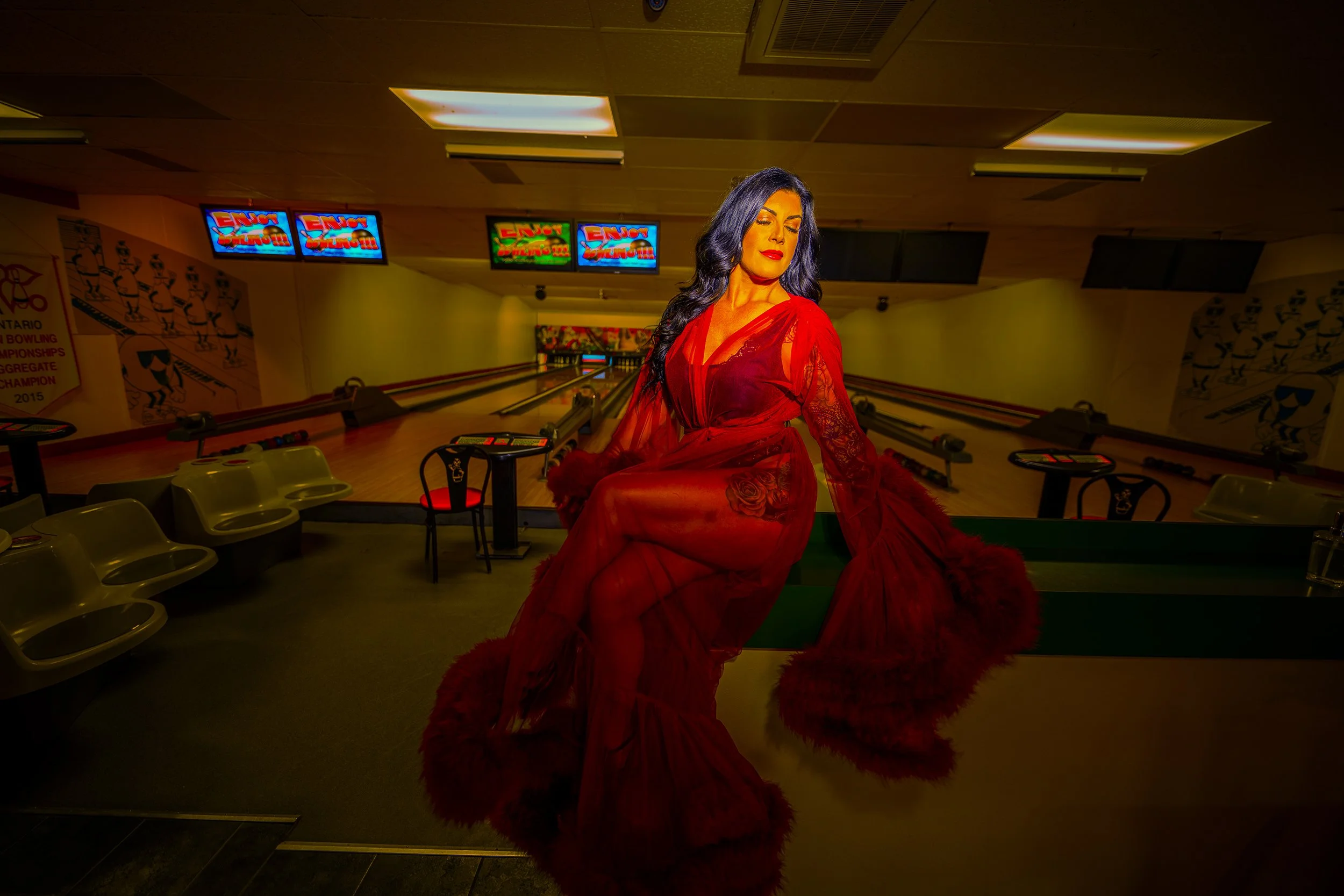 A woman with long dark hair and red lipstick, wearing a flowing red dress with floral patterns, sitting on a pool table in a bowling alley with multiple screens and bowling pins in the background.