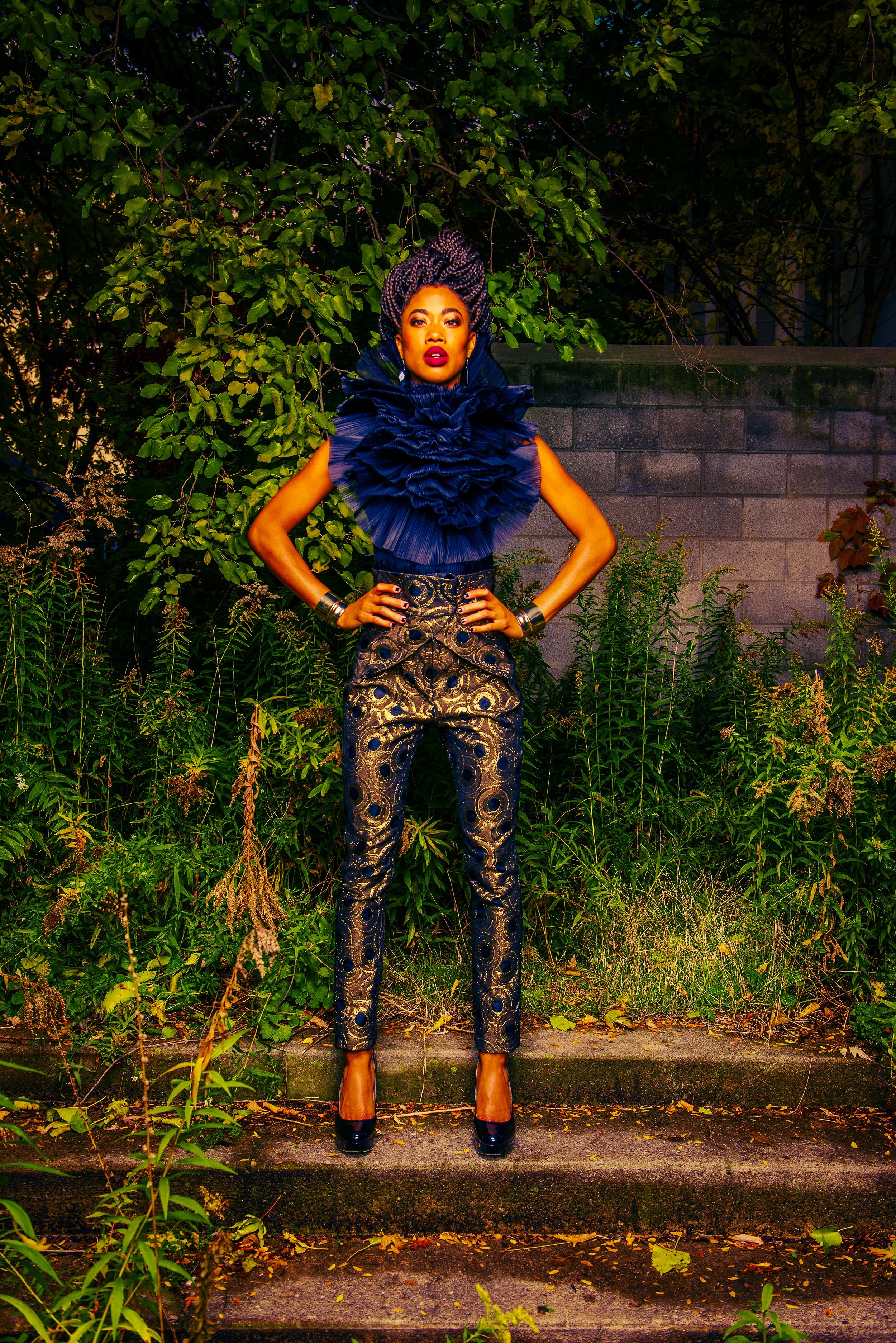 A woman standing outdoors at night, wearing an elaborate dark blue ruffled top and patterned pants, with her hands on her hips, in front of a green leafy hedge and a gray brick wall.