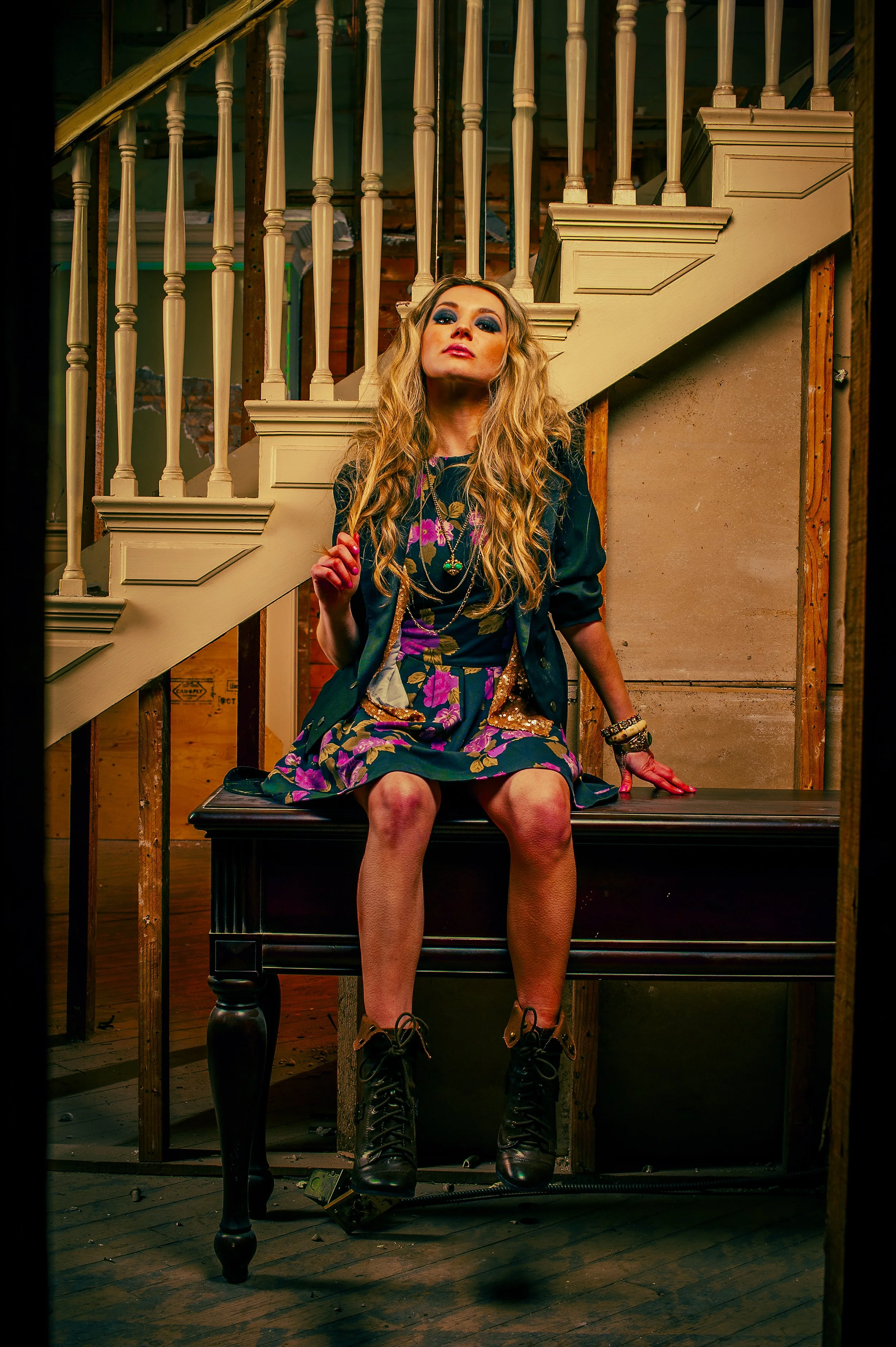 A woman with long blonde curly hair, wearing bold blue eye makeup, a floral dress, and black combat boots, sits on a black table in a partially unfinished room with exposed wooden studs and an ornate staircase railing behind her. She is looking confi