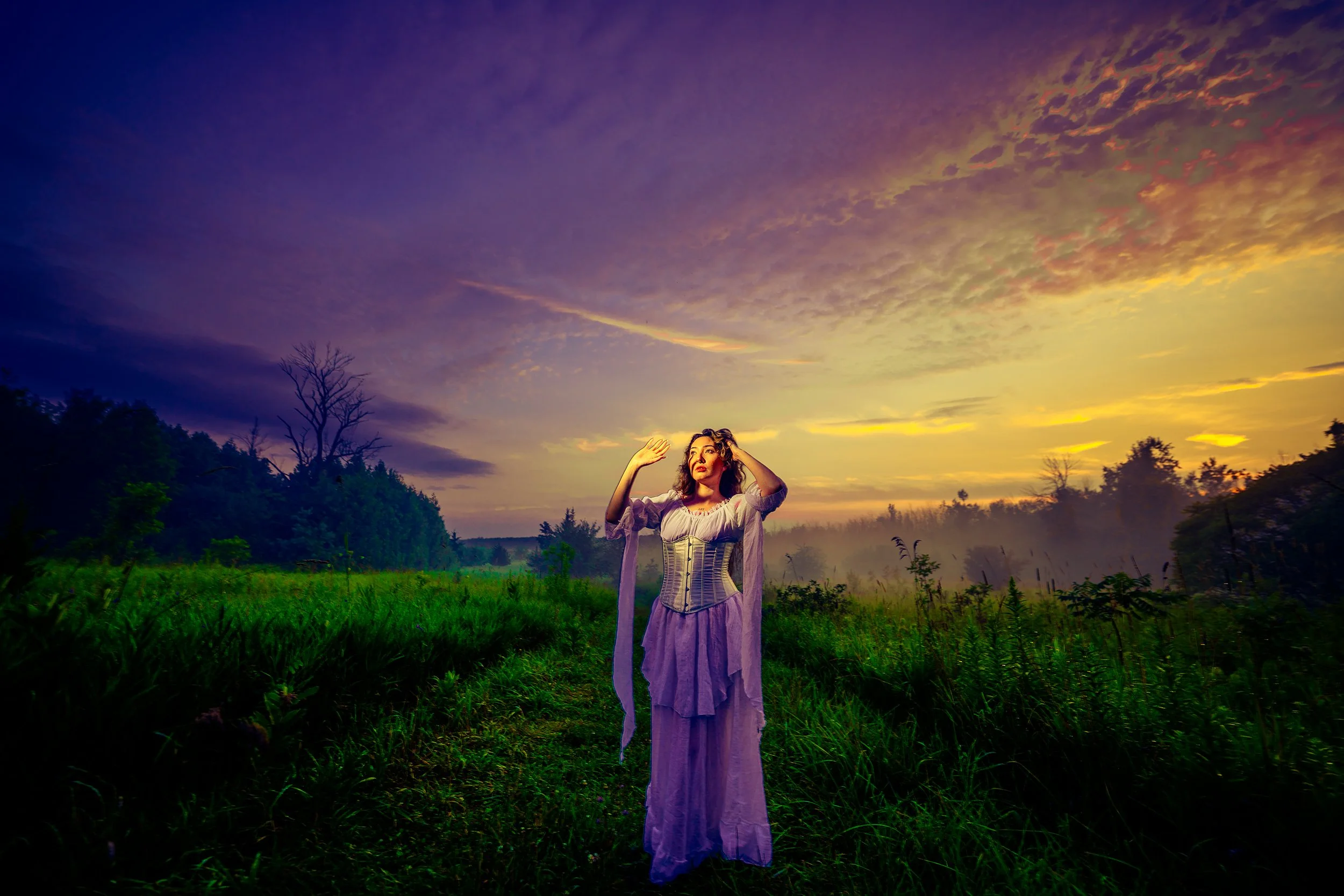 A woman in a flowing dress standing in a field during sunset, with her hands raised near her head and a colorful sky of purple, yellow, and orange in the background.