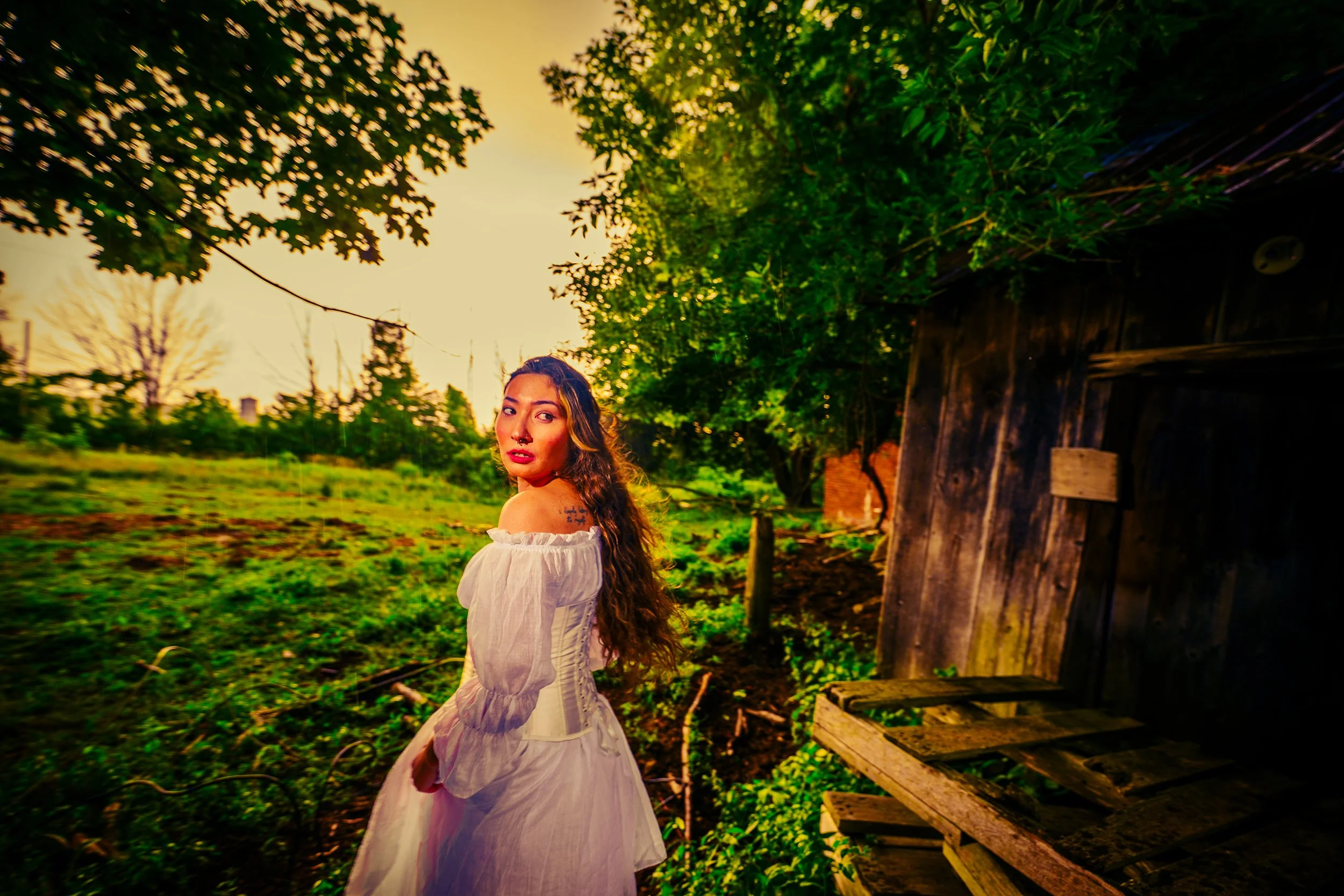 Young woman with long curly hair in a white dress standing outdoors near a wooden structure during sunset, surrounded by green trees and grass.