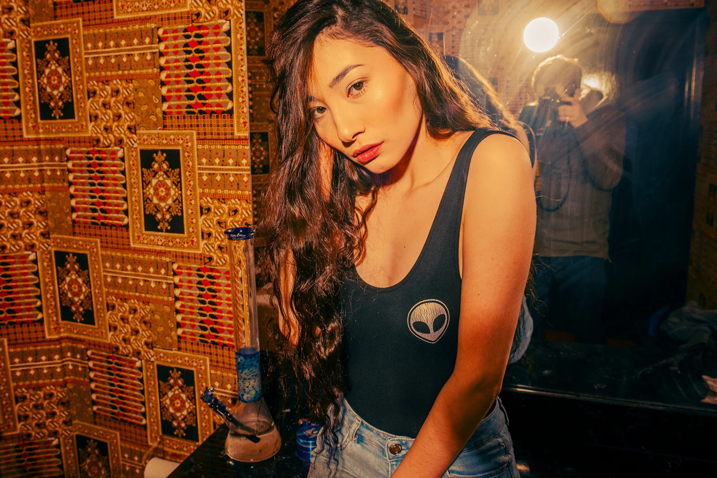 A young woman with long dark wavy hair, wearing a black tank top with an alien graphic, standing indoors near a display of colorful patterned textile or carpet. The background features warm lighting and a brick archway.